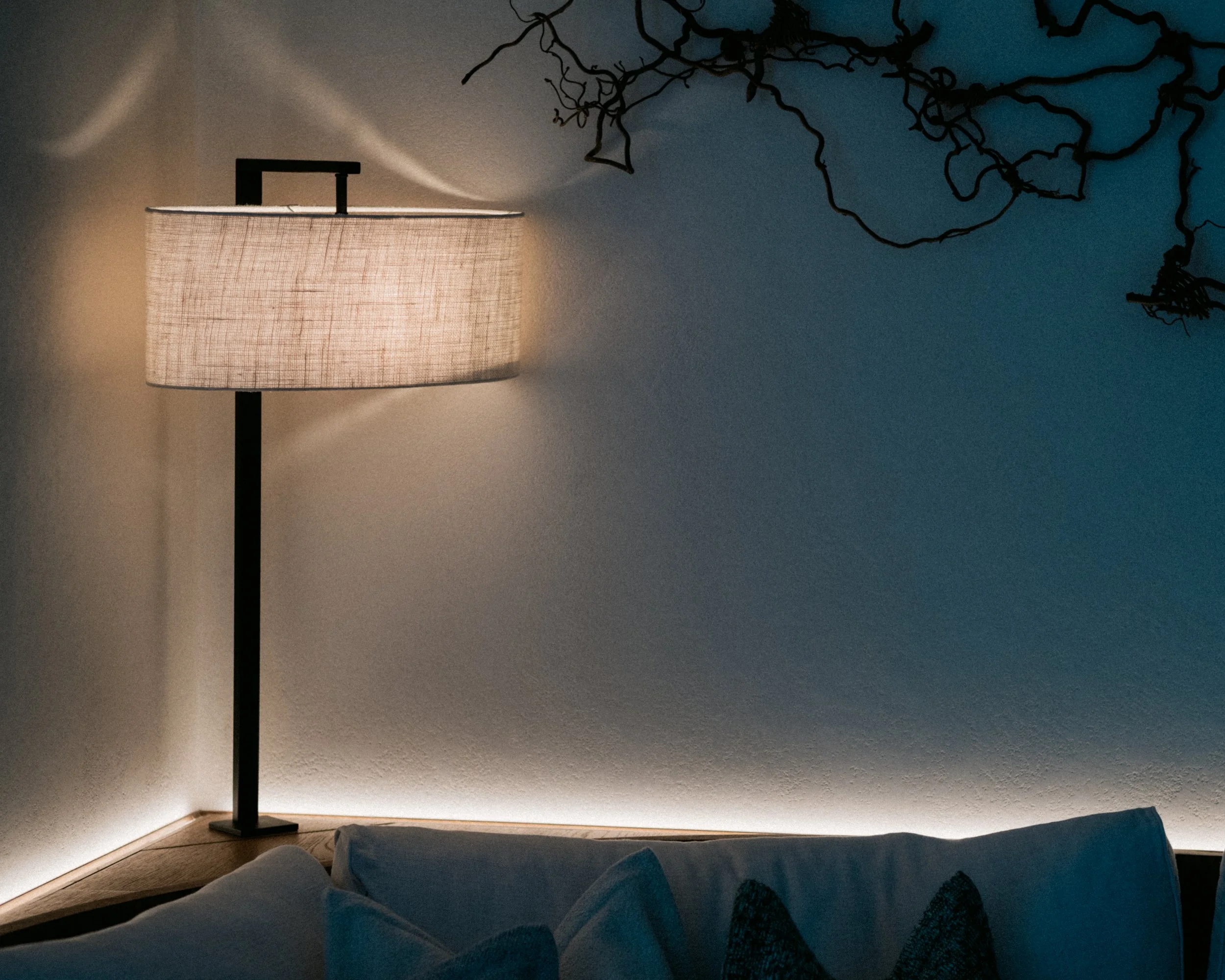 A corner of a room illuminated by a tall floor lamp with a circular fabric shade, casting light on the wall. There are dark, twisted tree branches or wires mounted on the wall, and part of a bed with a dark blanket and some pillows is visible in the 