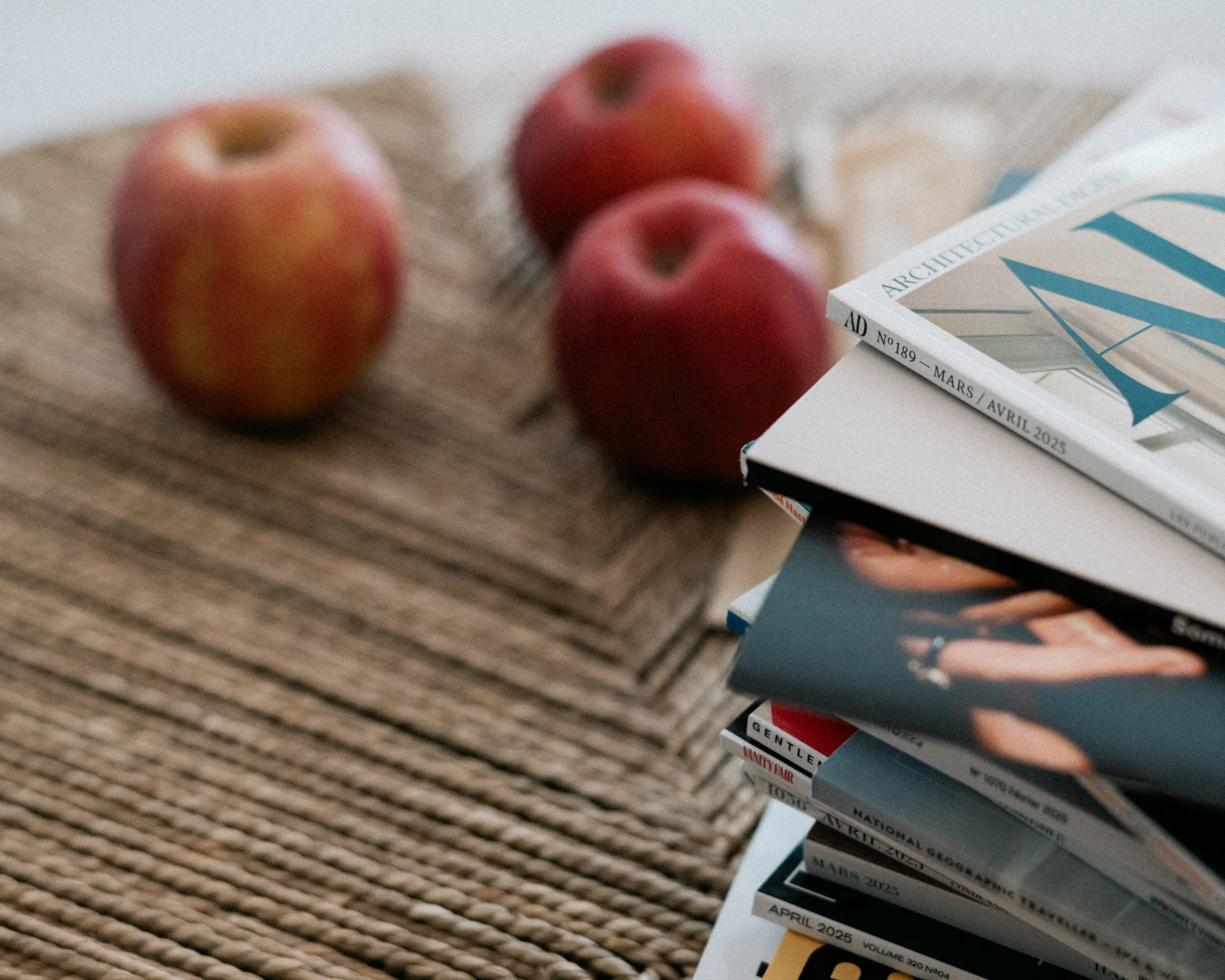 Close-up of three apples on a textured surface next to a stack of magazines or catalogs, with the focus blurred and no clear background.