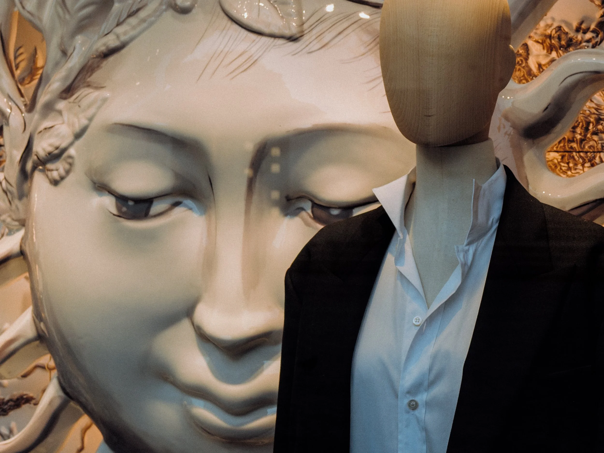 Close-up of a person wearing a white shirt and black blazer, with a partial view of a sensual, classical-style mask face sculpture in the foreground.