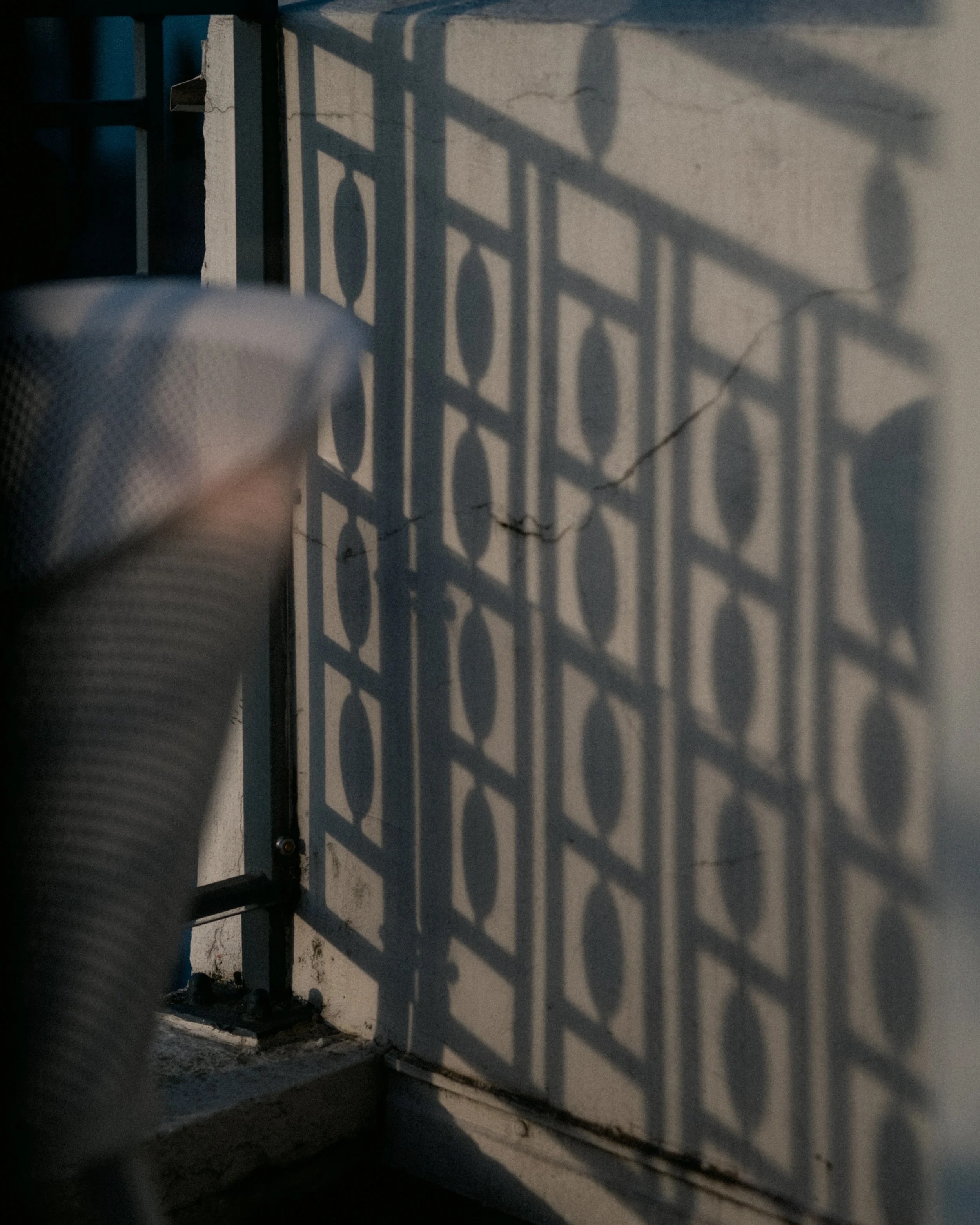 A shadow of a metal fence with decorative oval and square shapes on a wall, with part of a person's leg and shoe visible in the lower left corner.