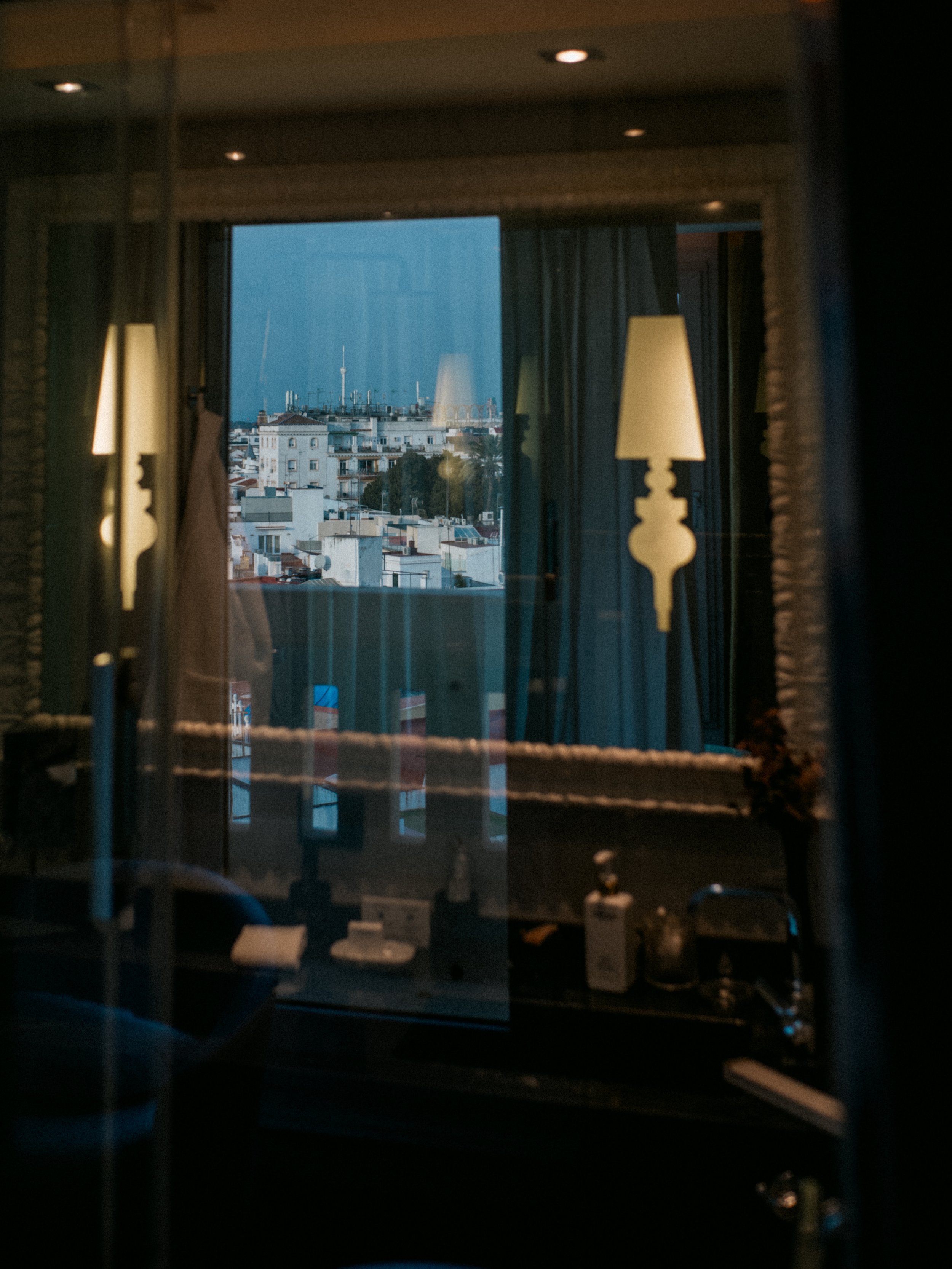 Cityscape with buildings viewed through a window with curtains and lamps inside a room.