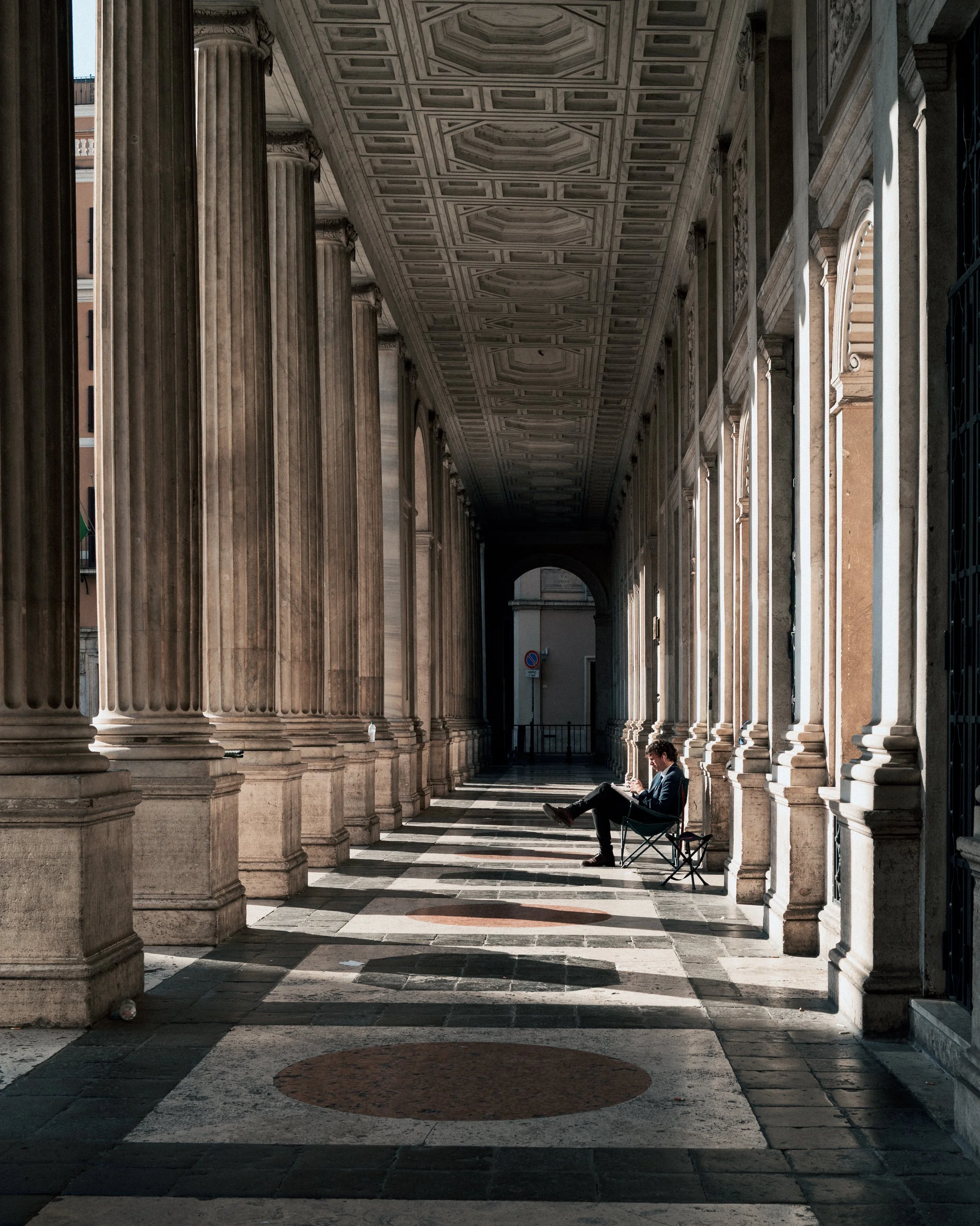 A man reading a book while sitting on a chair under a colonnade with large, classical columns and a decorated ceiling, casting shadows on the ground.