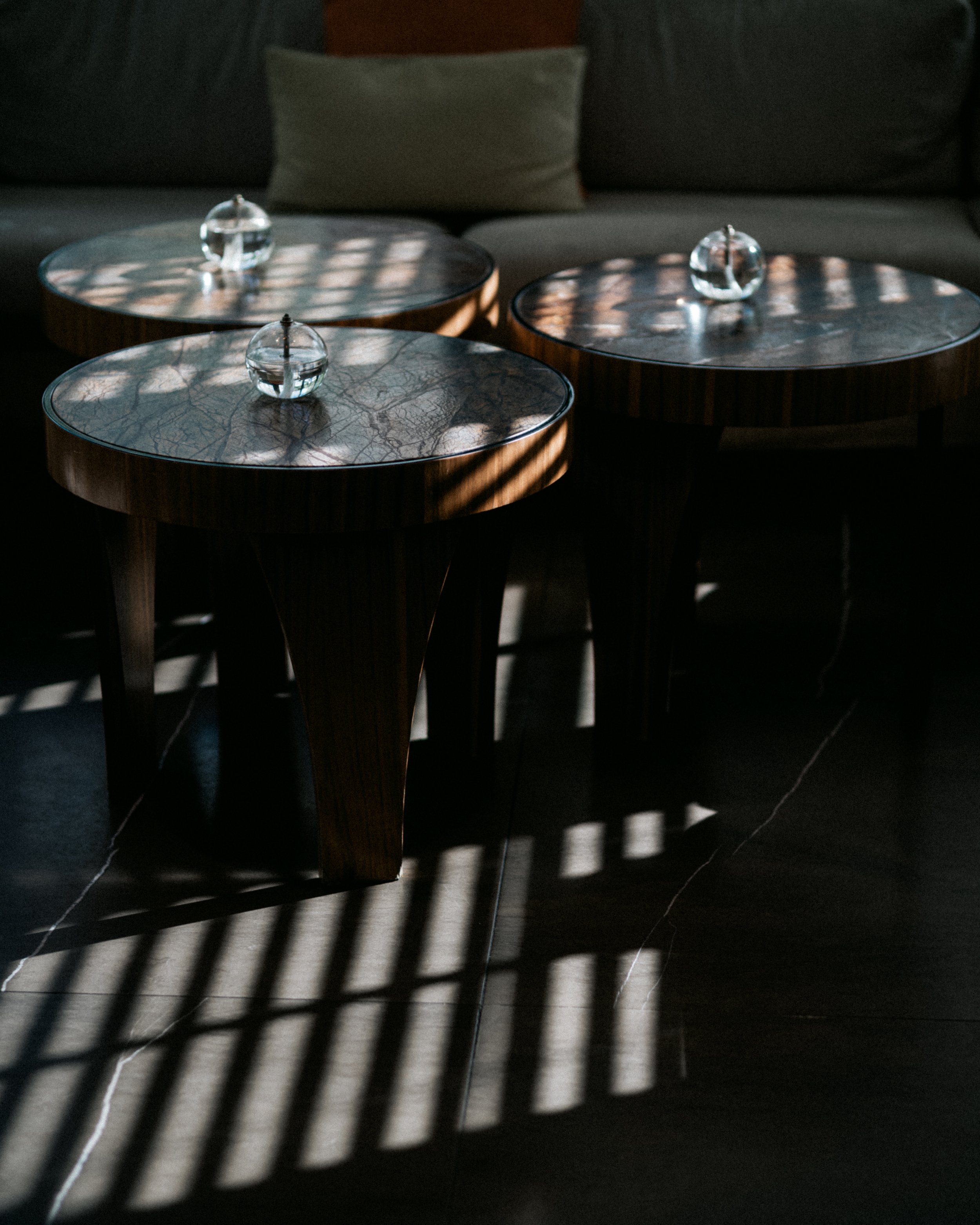 Three round tables with marble tops and glass decorative objects, casting striped shadows on a dark floor in a dimly lit room.