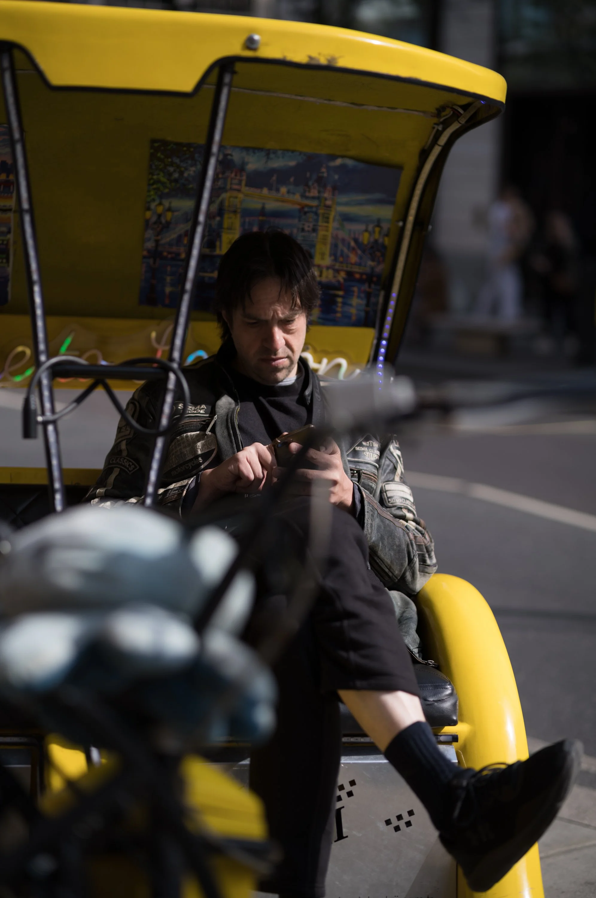 A man sitting on a yellow pedicab at night, looking at his phone, with a cityscape painting on the back of the pedicab.