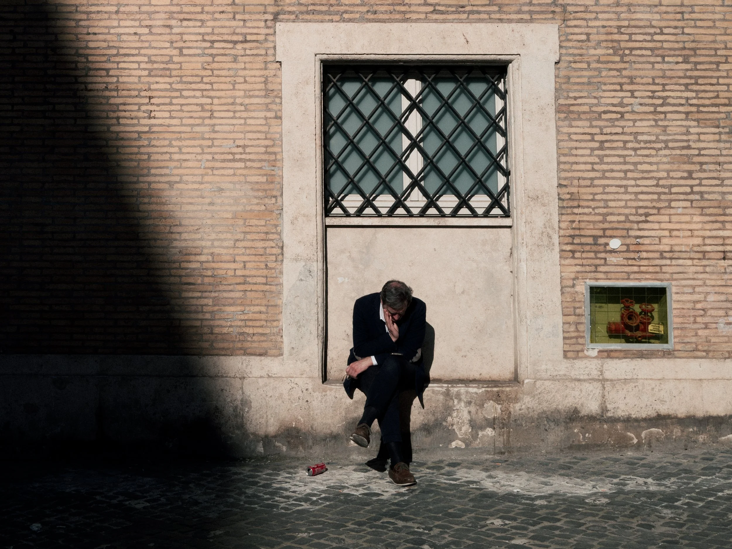 A man in a dark suit is crouching and holding his left knee, appearing injured, on a cobblestone street in front of a brick wall with a window. A red spray paint can is on the ground nearby.