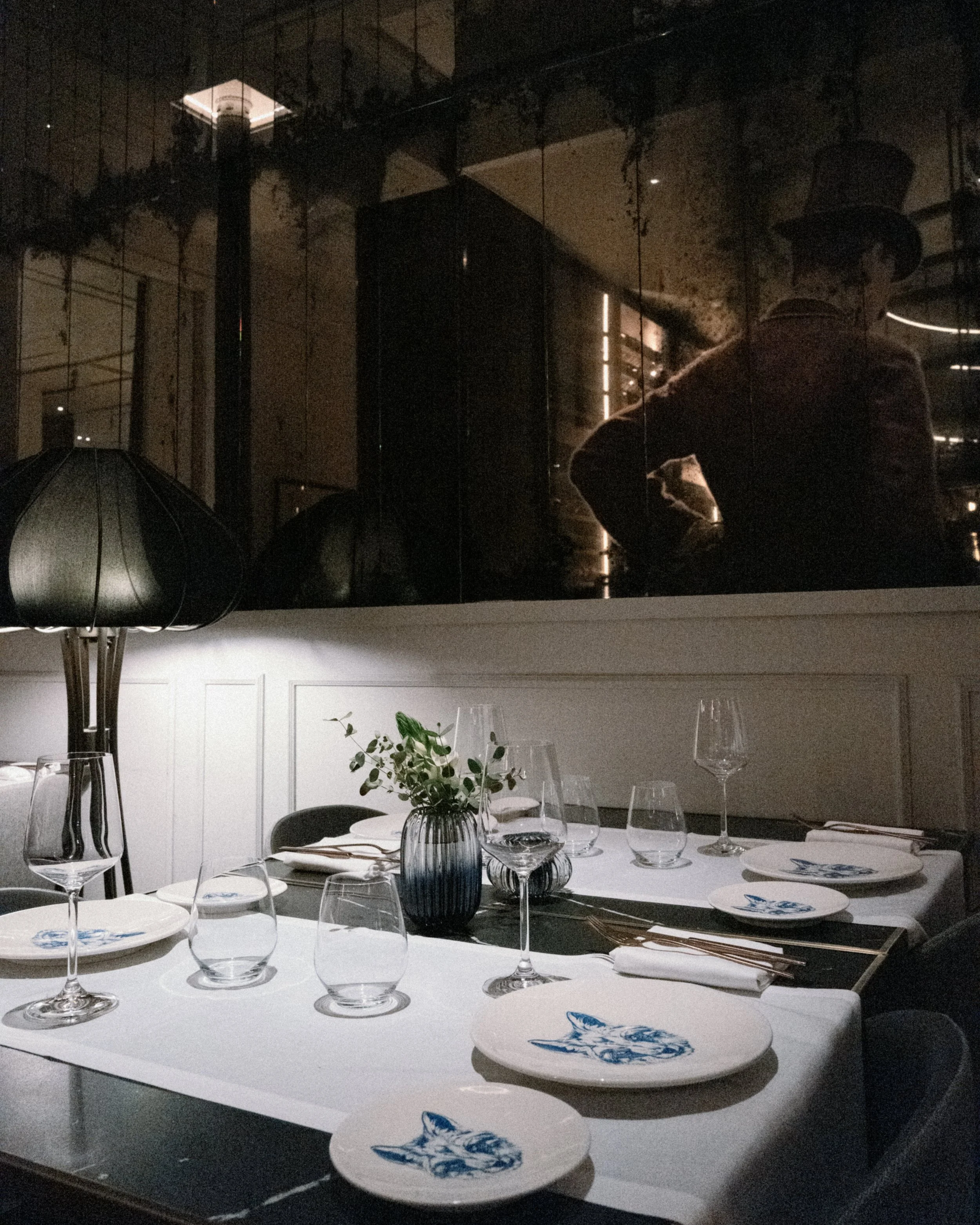 A formal dinner table set with empty wine glasses, plates, and cutlery, decorated with a dark blue flower-patterned ceramic tray and a small vase with green foliage. The background shows a dark, reflective window or wall with a person's reflection we