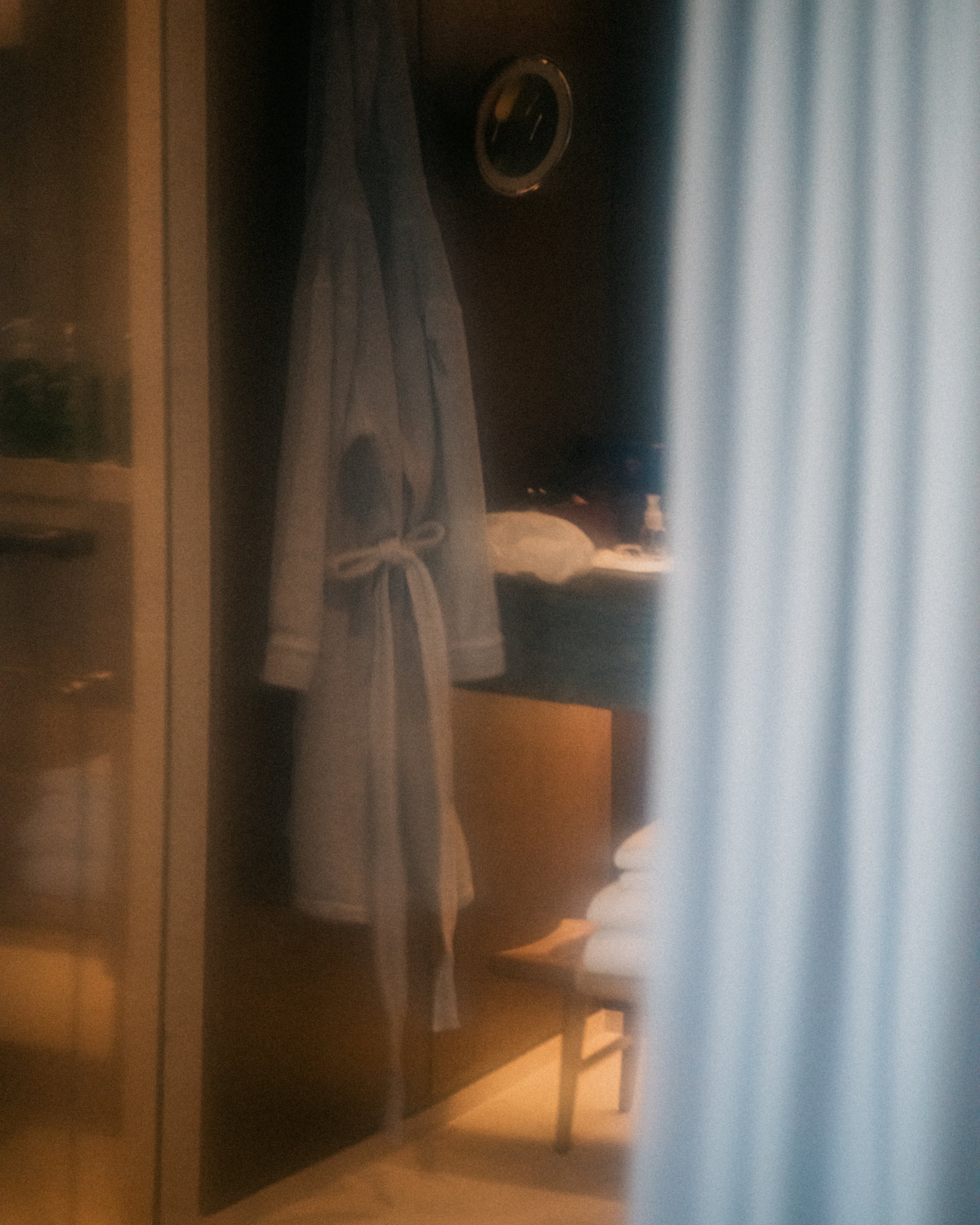 A wooden nightstand with a round alarm clock on top, a folded gray towel, and some items underneath, partially visible. A blue curtain is in the background.