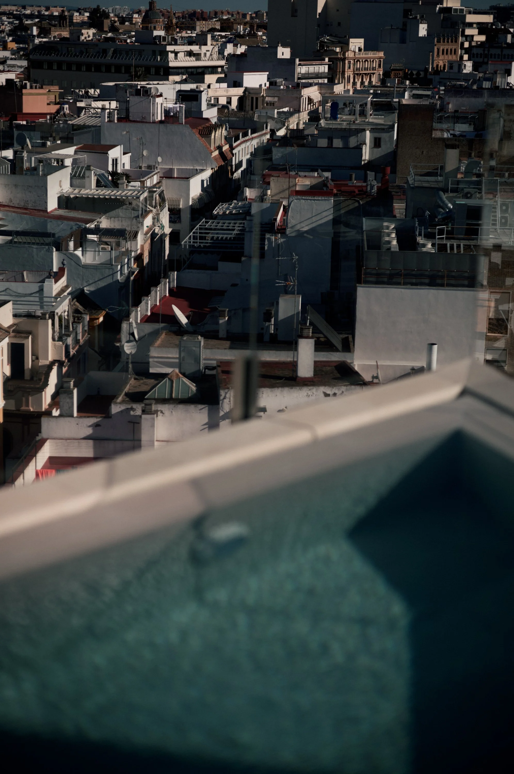 A high-angle view of a city skyline with densely packed buildings, some with solar panels, antennas, and satellite dishes, seen through a glass window.