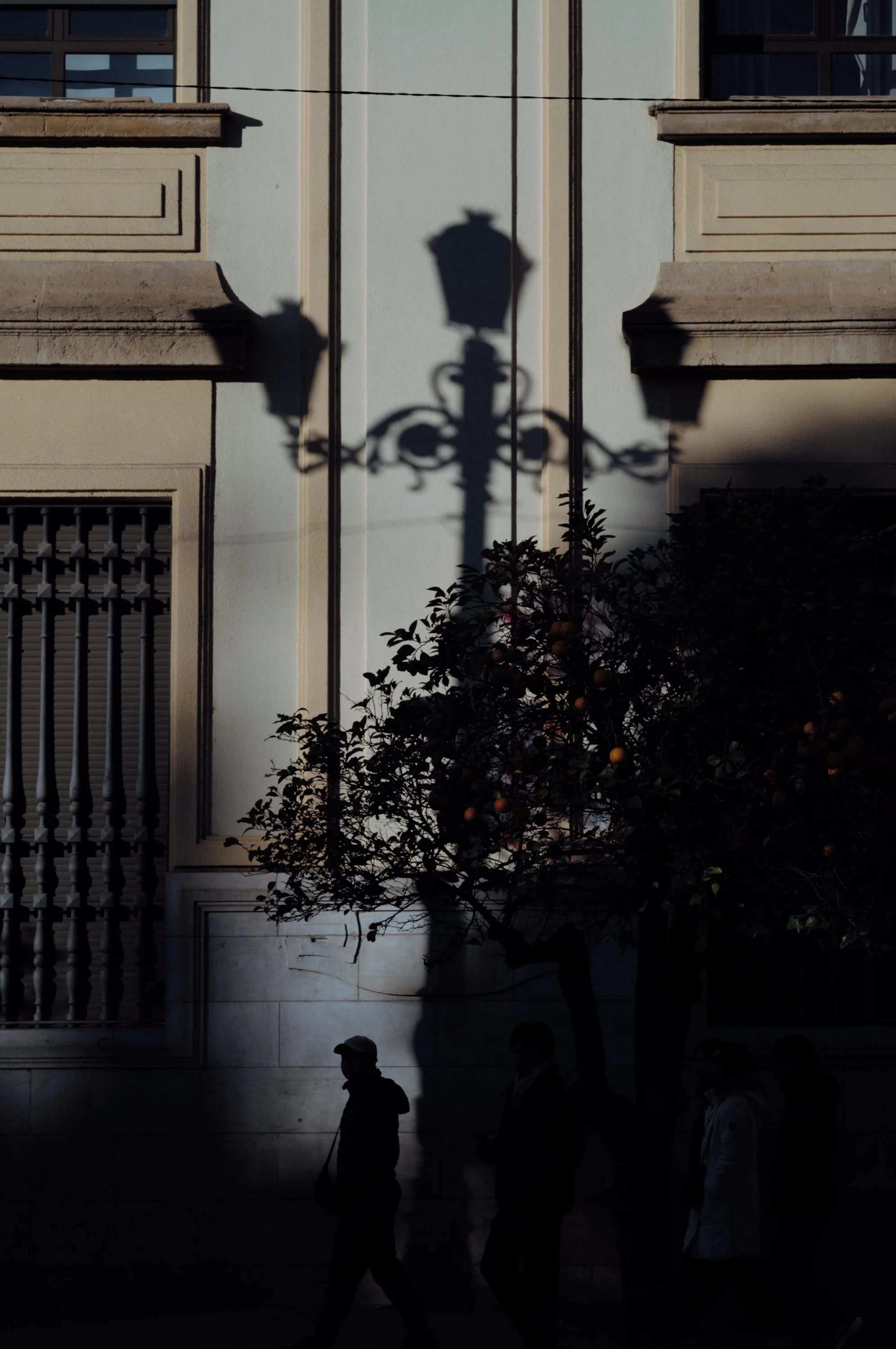 Shadow of a vintage streetlamp cast on the side of a building with three pedestrians walking past and a fruit tree in front.