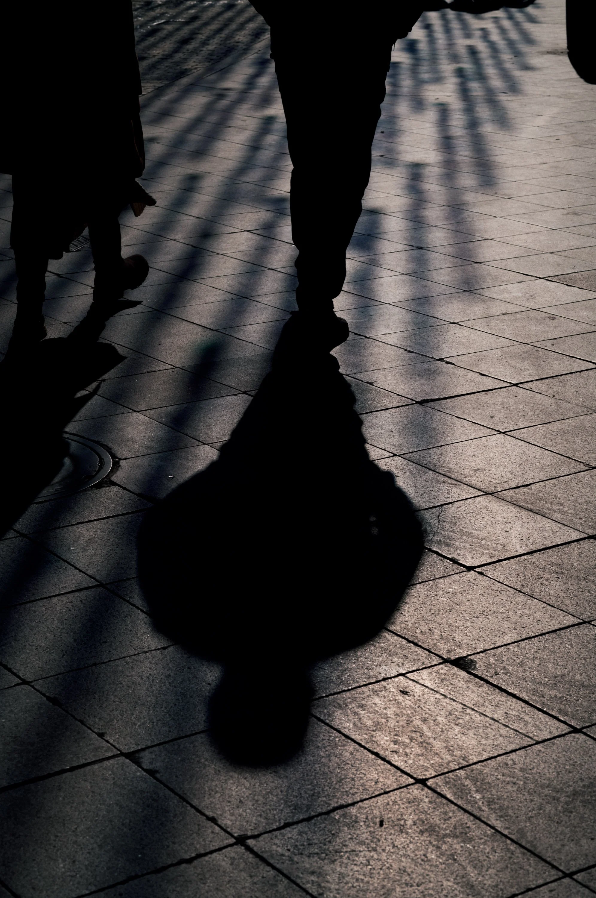 Silhouette of a person walking with a bag, cast long shadow on a tiled sidewalk, with shadows of railings and other pedestrians.