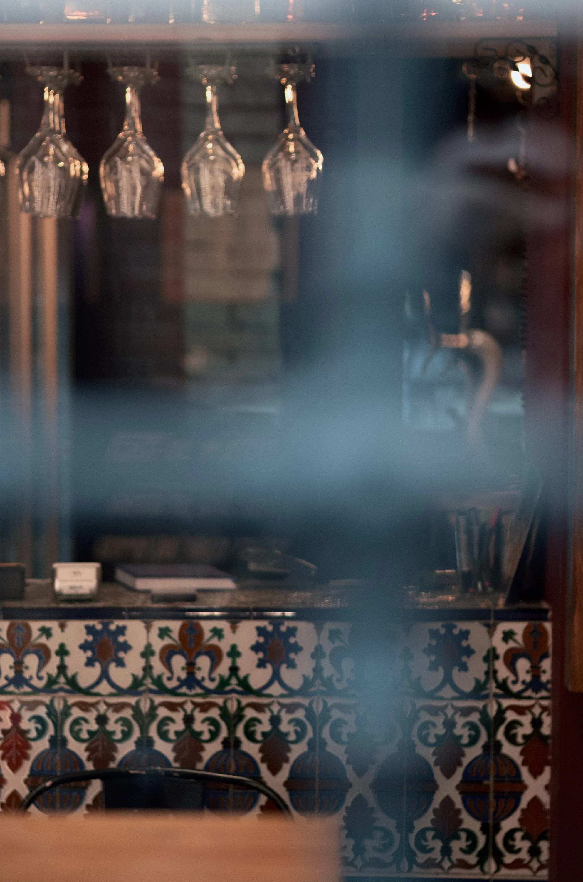 Blurred glasses hanging upside down above a bar counter with books and a decorative tiled wall in the background.