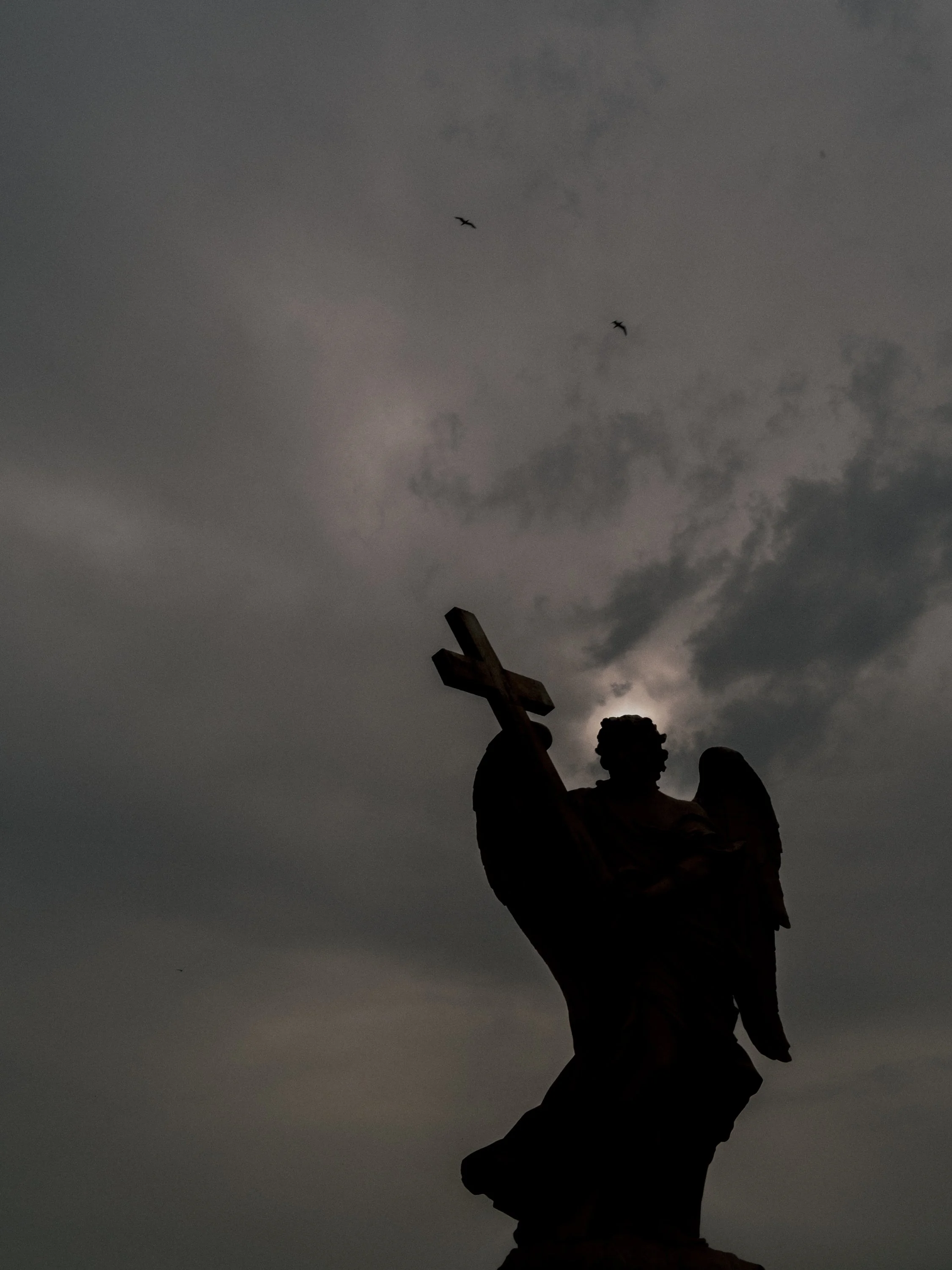 Silhouette of a statue of an angel holding a cross against a cloudy, dusk sky with two birds flying nearby.