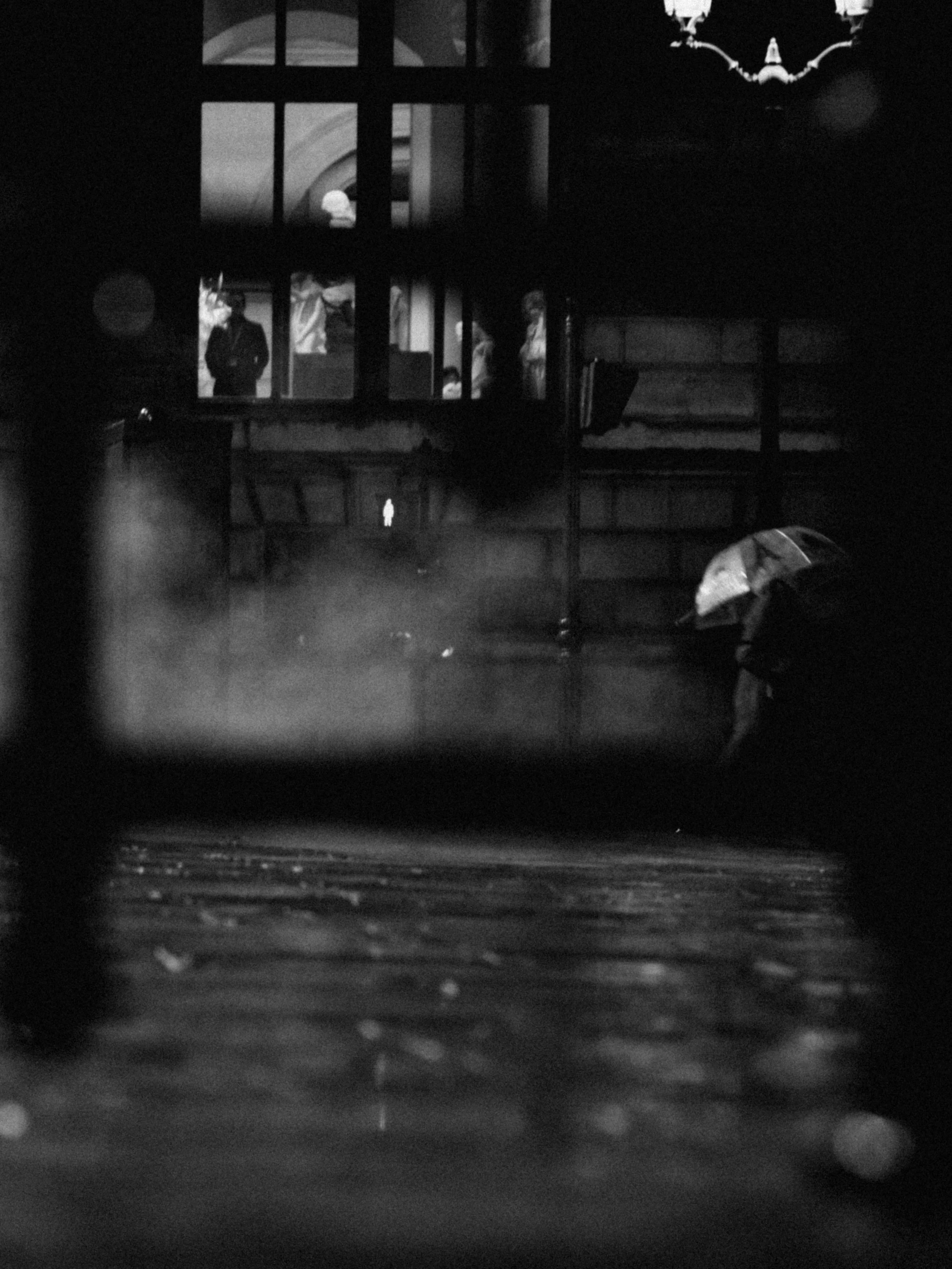 A black and white photo of a person walking with an umbrella in the rain, seen through a window.