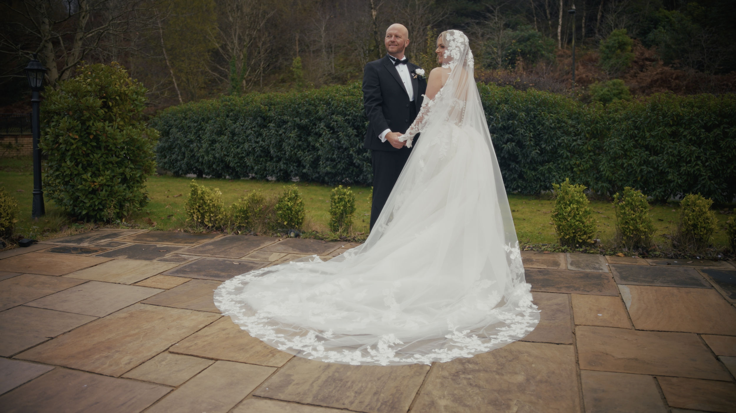 A bride and groom standing outdoors on a stone patio, holding hands and looking at each other, with trees and bushes in the background. The bride is wearing a white wedding dress with a long train and lace veil, while the groom is dressed in a black 