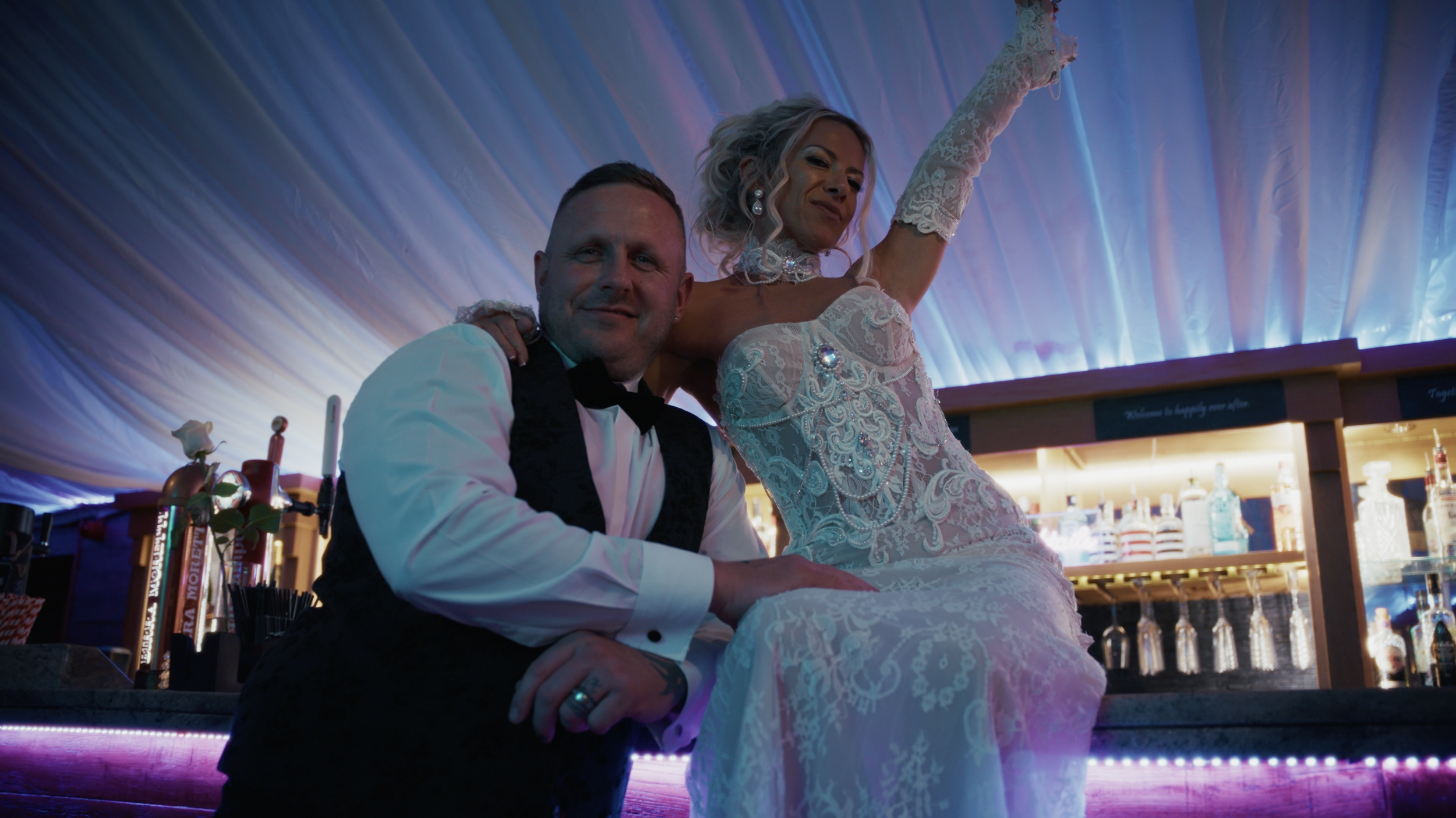 A newlywed couple, the groom in a black tuxedo and the bride in a white lace wedding dress, are seated together at a bar with a colorful, lit-up background, celebrating their wedding.