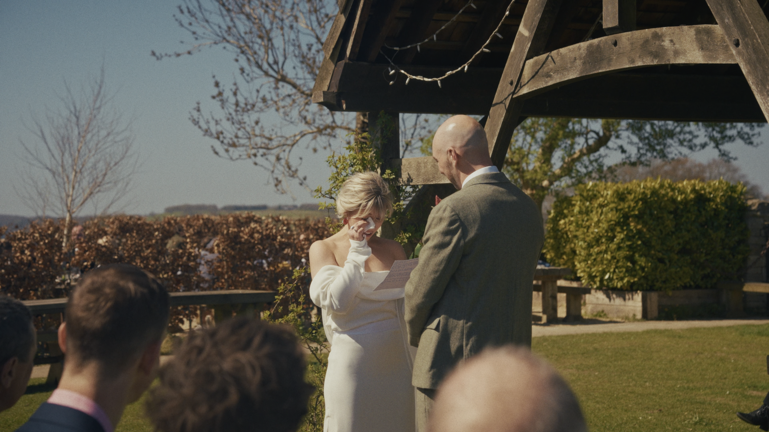 A woman in a white off-shoulder dress is crying while reading from a paper to a man in a gray suit at an outdoor wedding ceremony on a sunny day, with guests seated in front and a wooden structure and trees in the background.