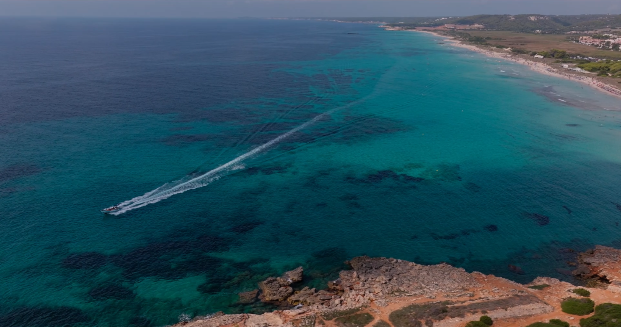 A boat speeding through turquoise waters near a rocky coastline and a sandy beach with buildings and greenery in the background.