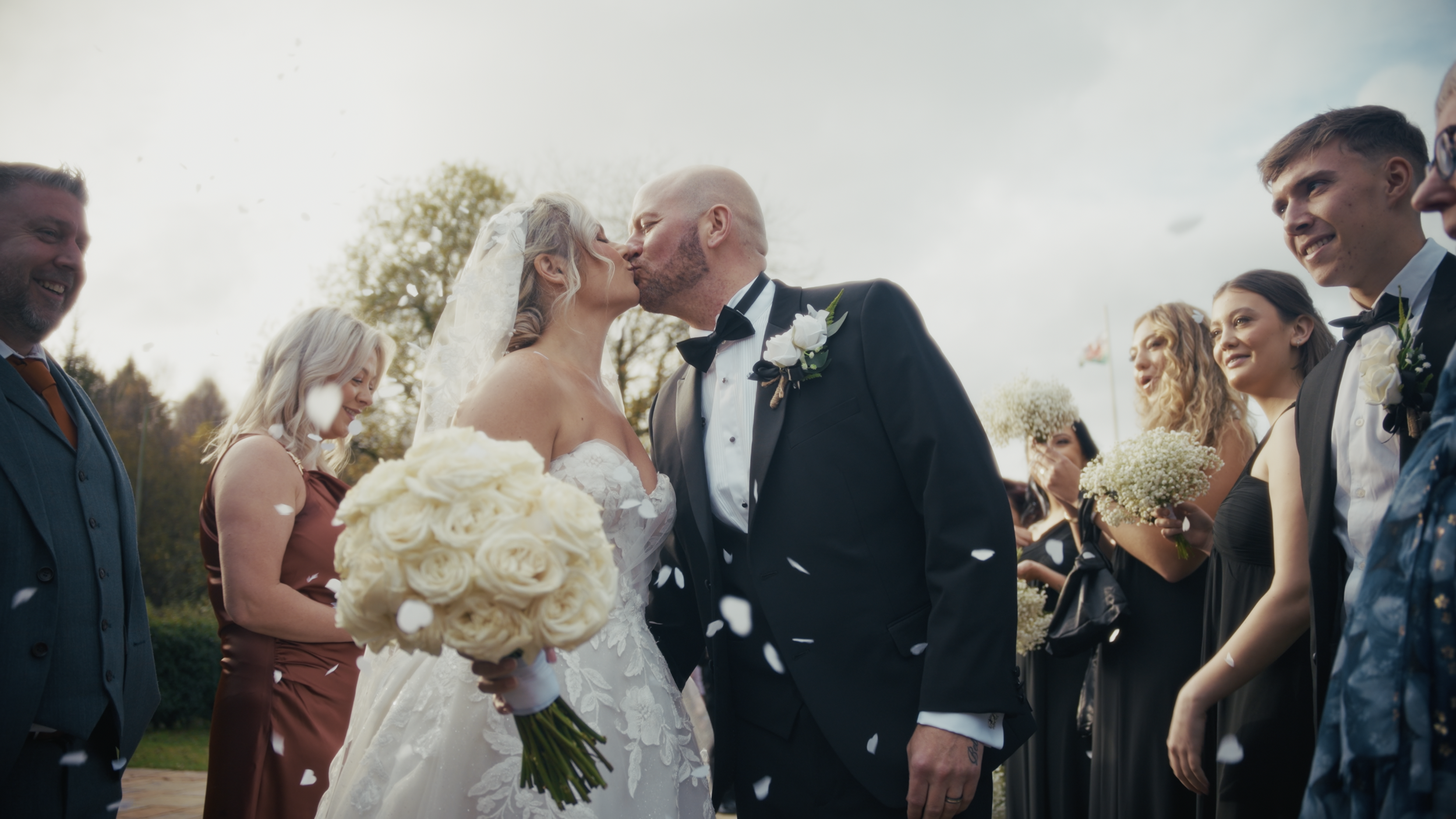A bride and groom kiss during their wedding ceremony, surrounded by their wedding party and guests, with white confetti falling.