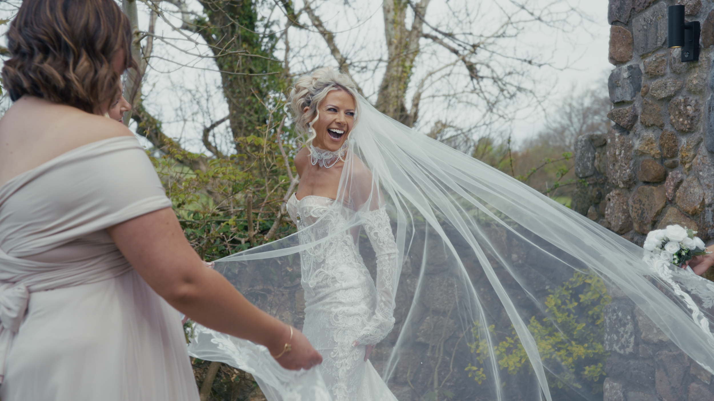 A joyful bride in a white wedding dress, with a lace neckline and long lace gloves, laughing outdoors near a stone wall, as her wedding veil is held by someone else.
