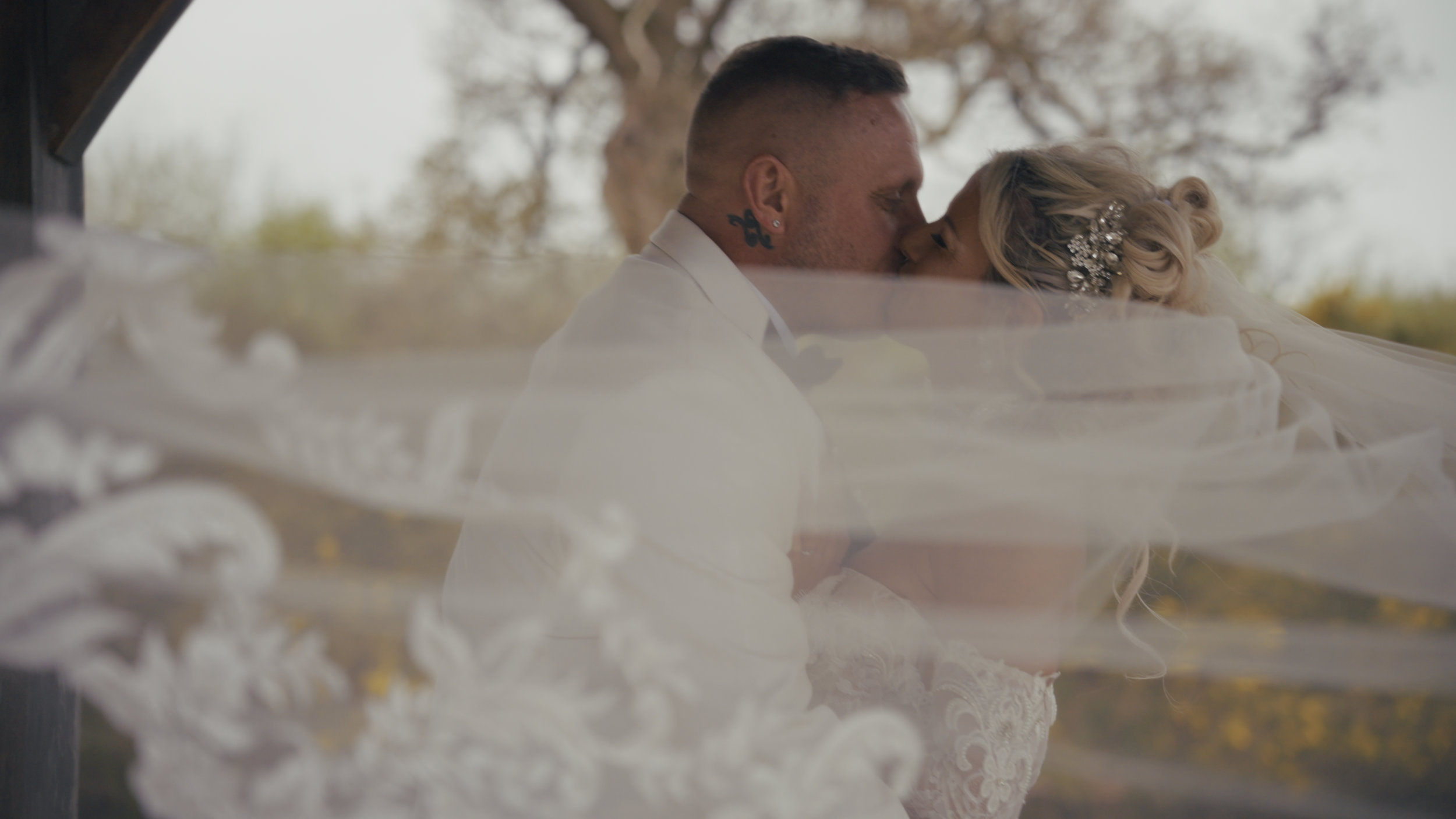 A bride and groom kissing on their wedding day, with the bride's veil partially in the foreground and trees in the background.