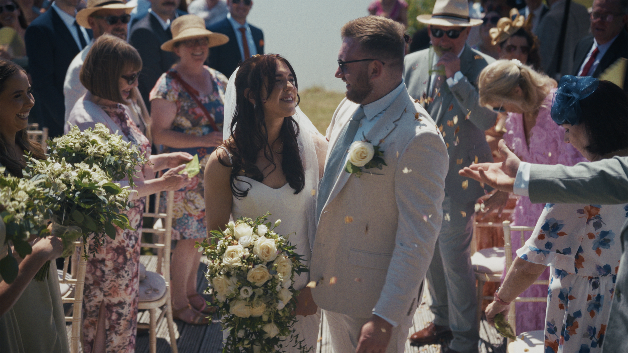 A bride and groom smile at each other during their outdoor wedding, surrounded by guests celebrating with flower bouquets and confetti.