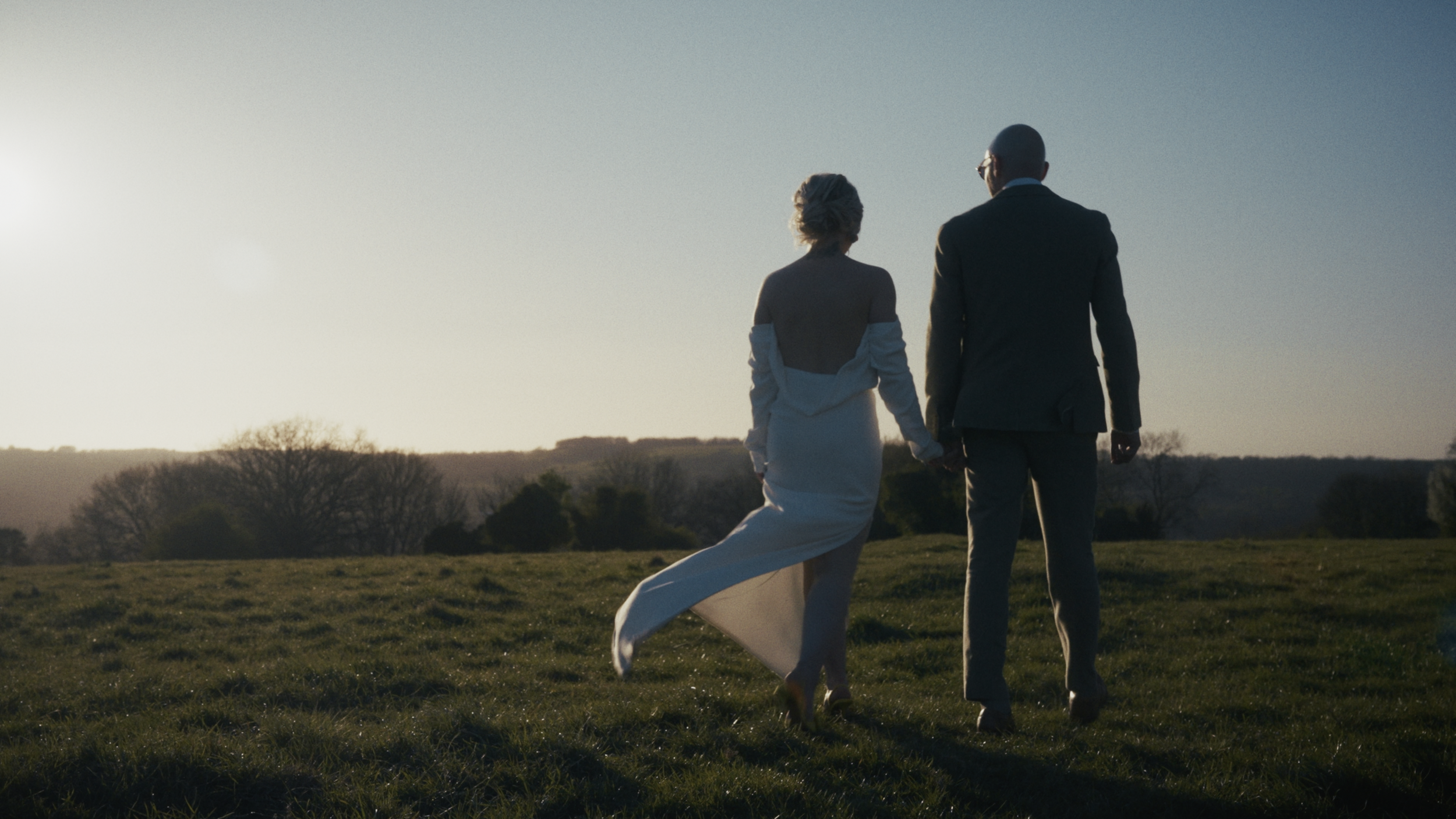 A couple walking hand in hand across a grassy field during sunset, with the woman in a white dress and the man in a dark suit.