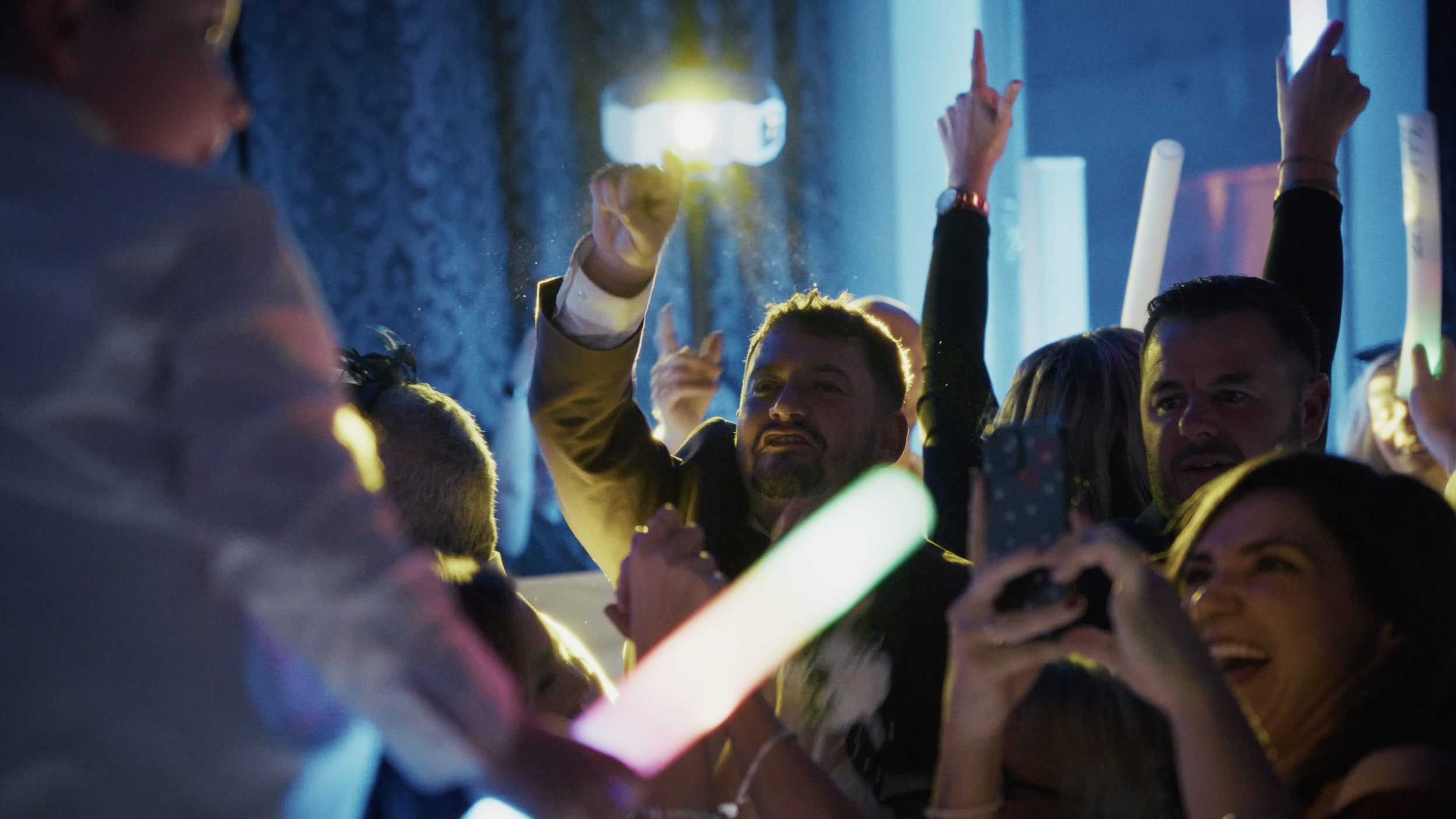 People at a party dancing and having fun, with some holding glow sticks and a person in the foreground taking a photo or video.