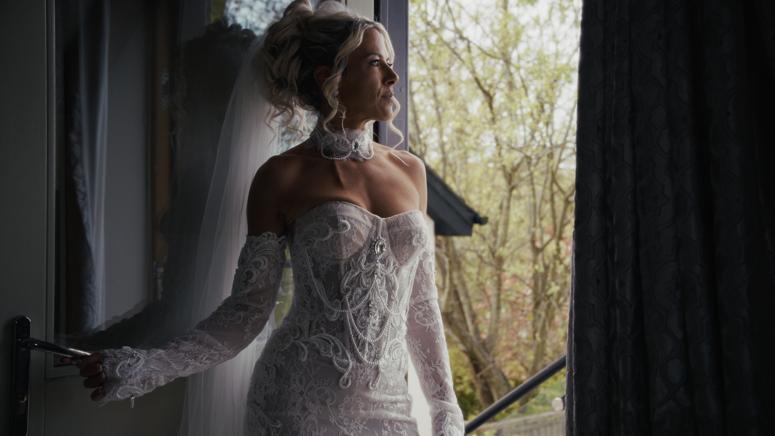 A bride in a lace wedding dress with long sleeves and an off-the-shoulder design, standing by a window, looking outside with a contemplative expression. She has blonde curly hair and is wearing a veil and a choker necklace.