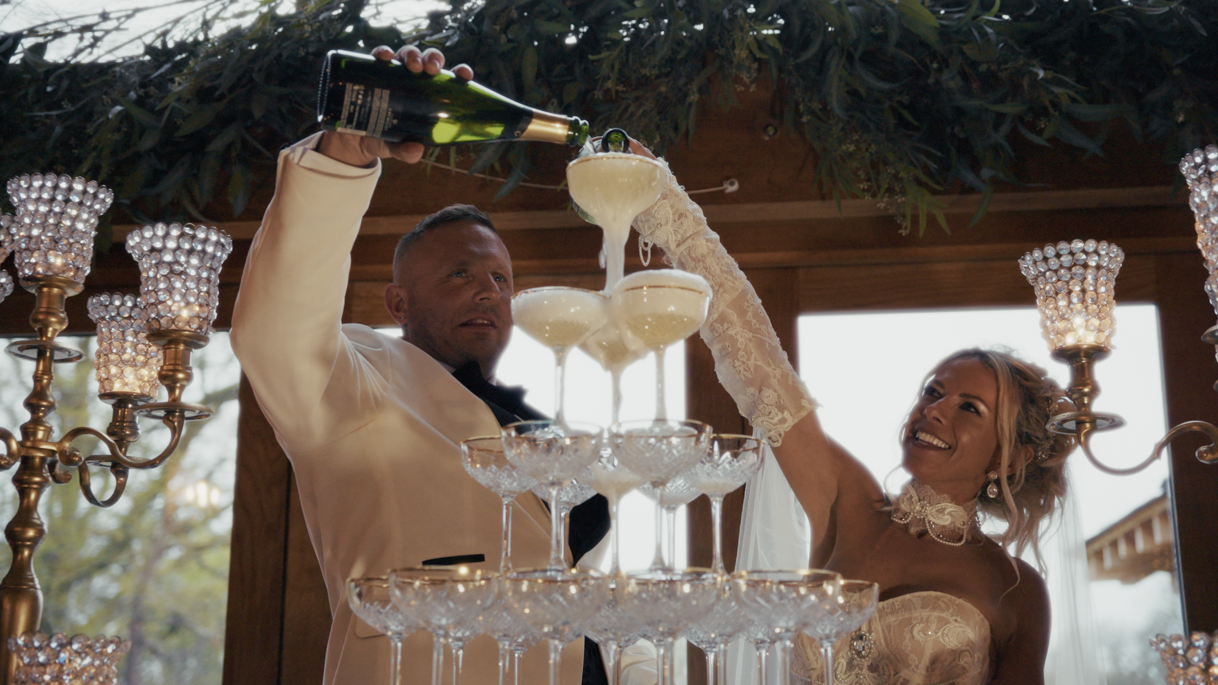 A couple at a wedding drinks champagne from a pyramid of glasses, with the groom pouring champagne and the bride smiling. They are inside a decorated venue, with crystal candle holders on chandeliers surrounding them.