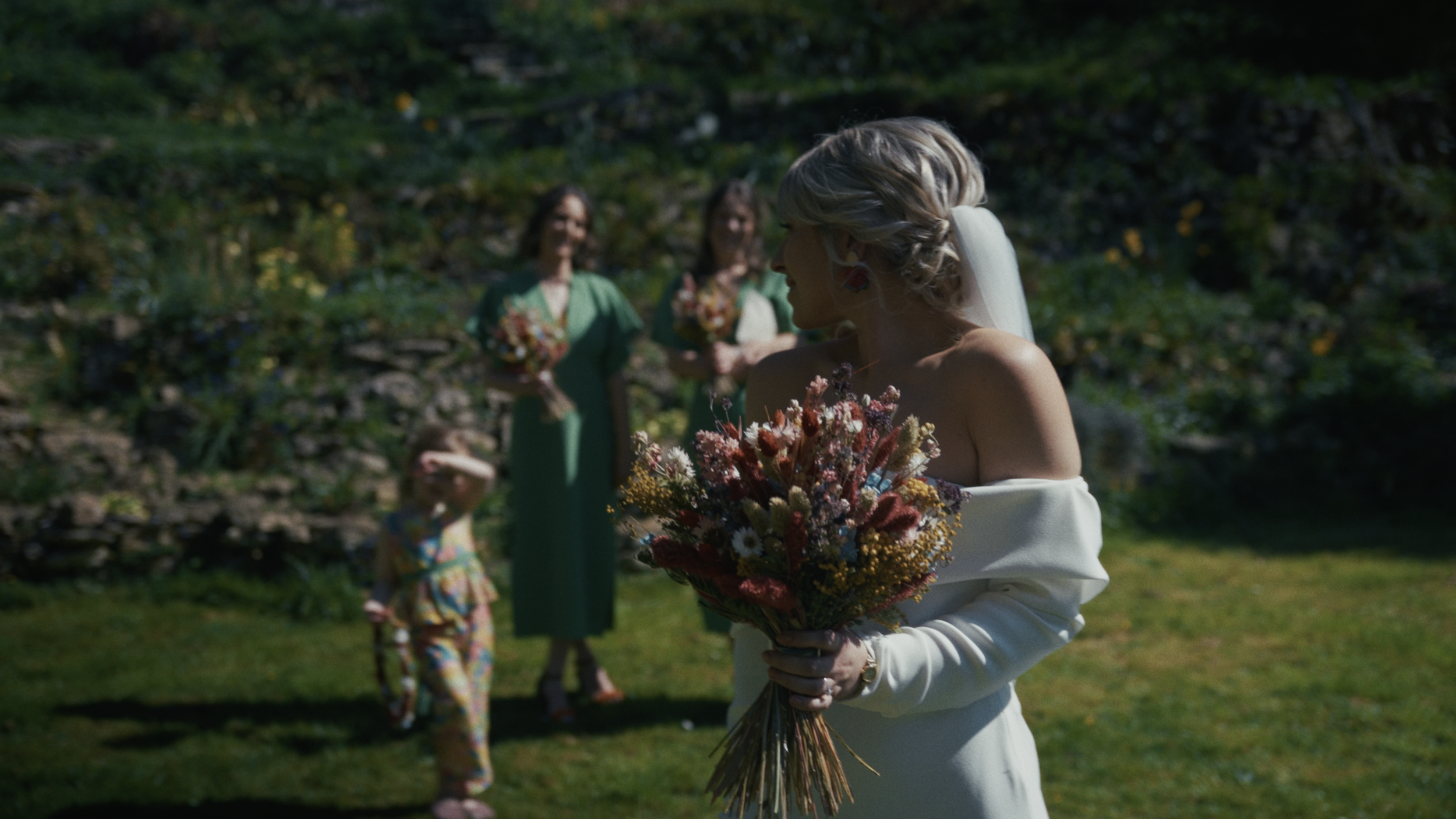A bride in a white off-shoulder dress holding a bouquet of flowers, standing outdoors with three women and a young girl in the background, on a grassy area with a stone wall and greenery.
