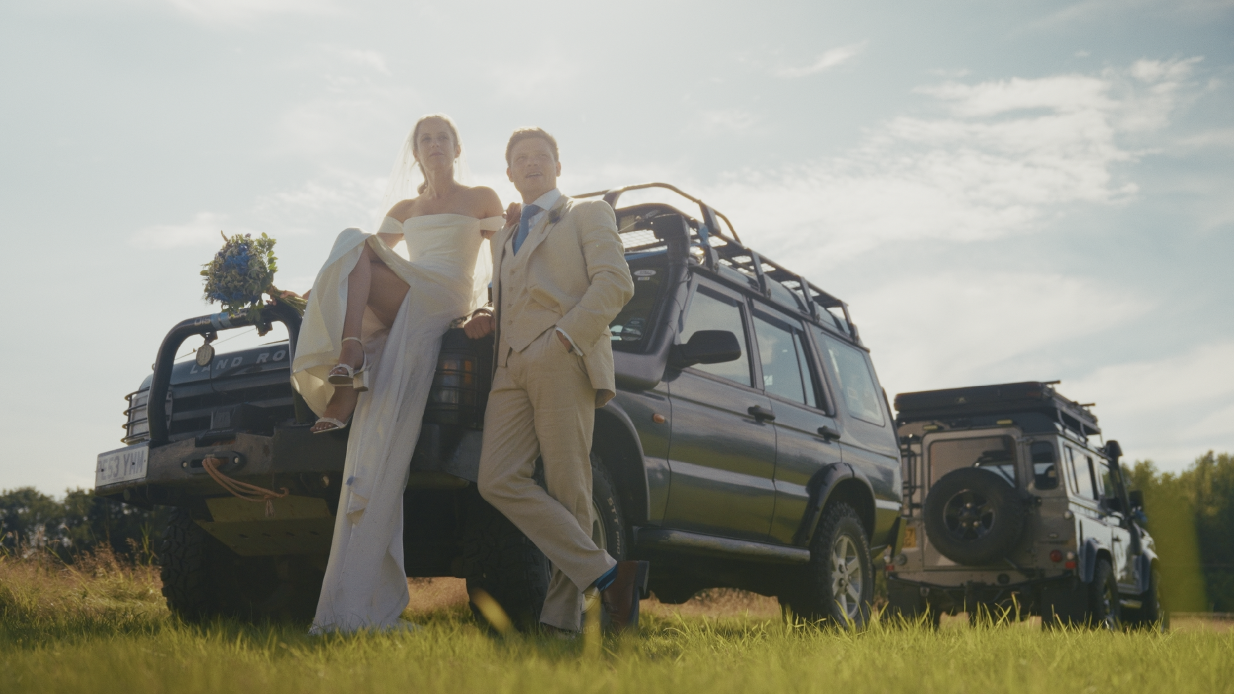 A bride and groom dressed in wedding attire sitting on the back of a Land Rover vehicle in an outdoor field, with a second vehicle behind them, both under a partly cloudy sky during daytime.
