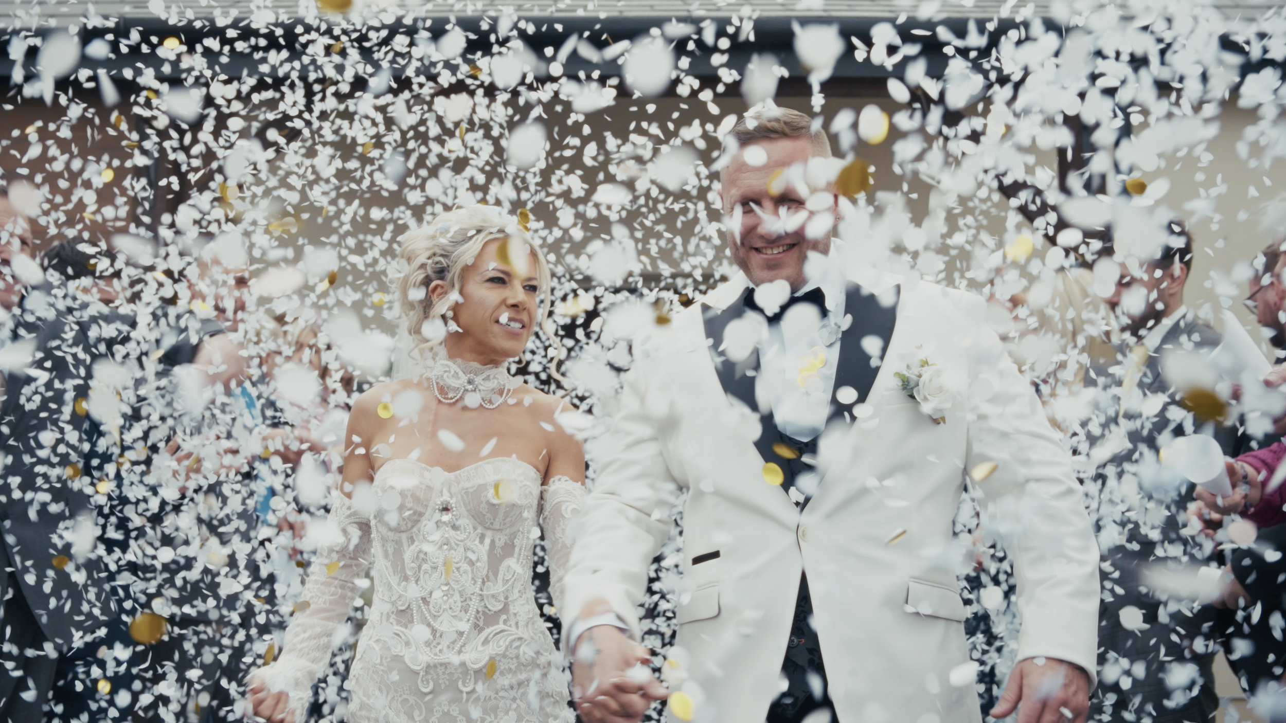A newlywed couple holding hands walking through falling confetti, surrounded by guests celebrating.