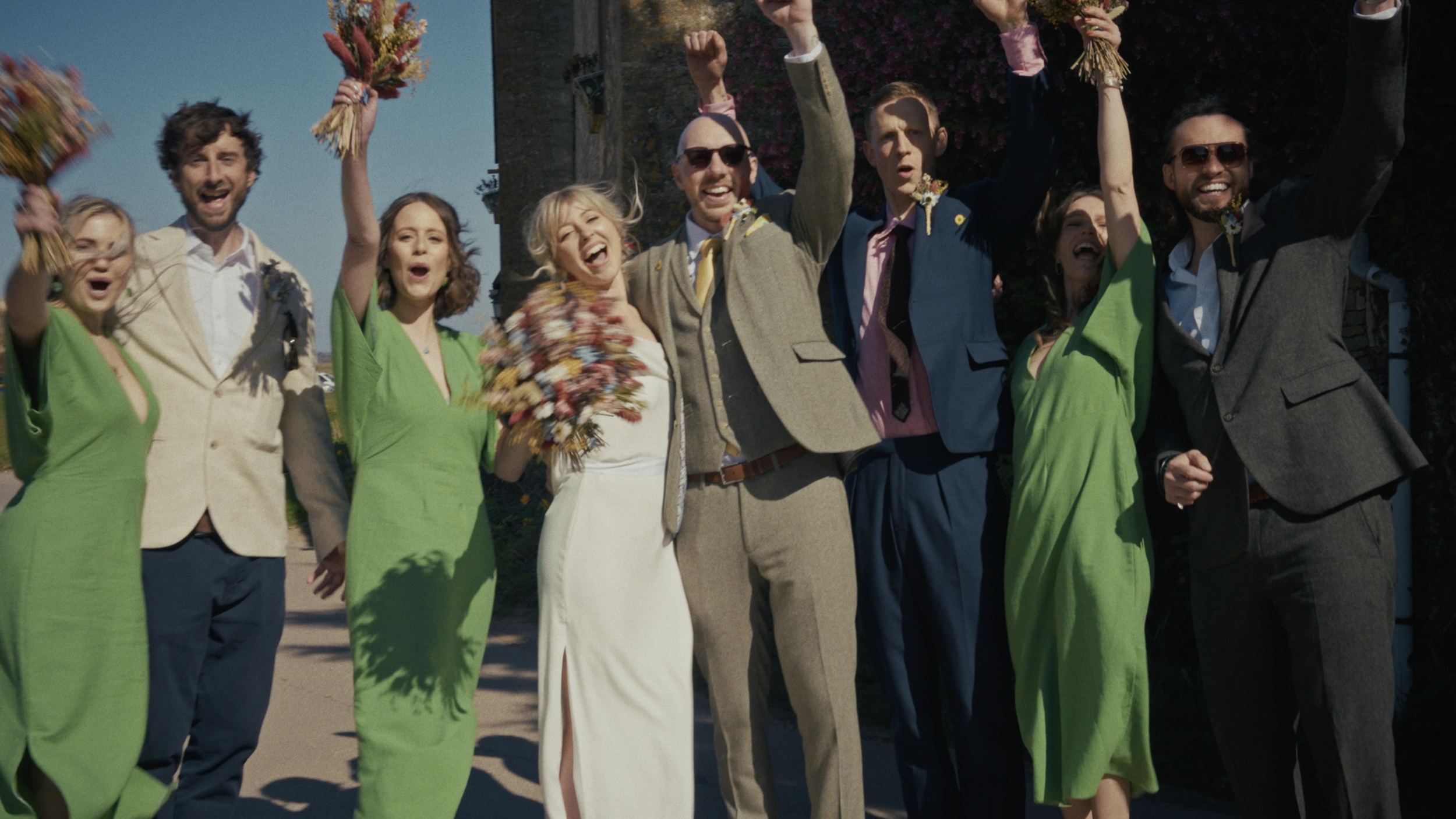 A group of people celebrating outdoors, wearing colorful attire, some holding flower bouquets, with happy expressions and raised fists.