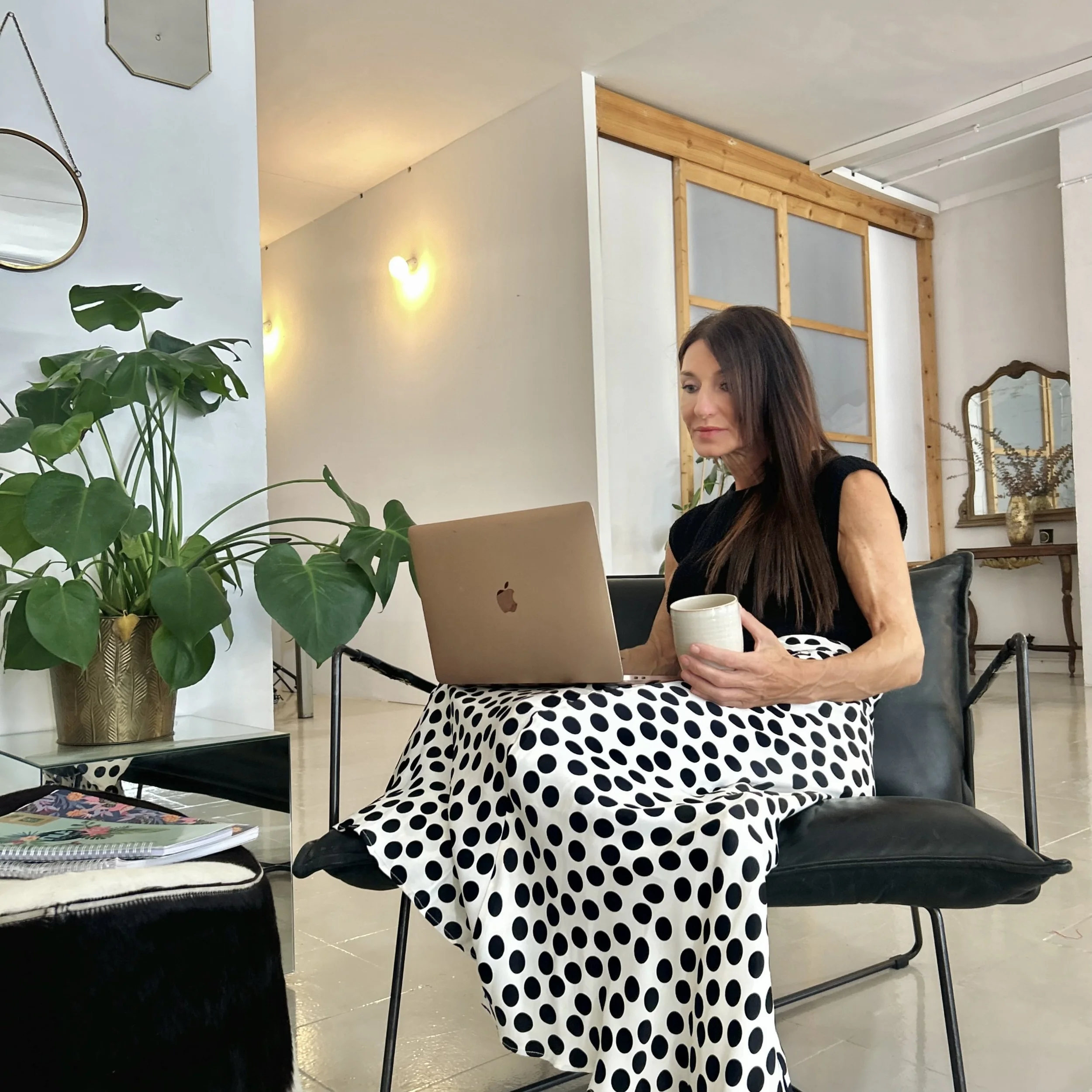 A woman with long brown hair sitting on a black chair, working on a laptop and holding a cup, in a modern interior space with a green plant nearby.