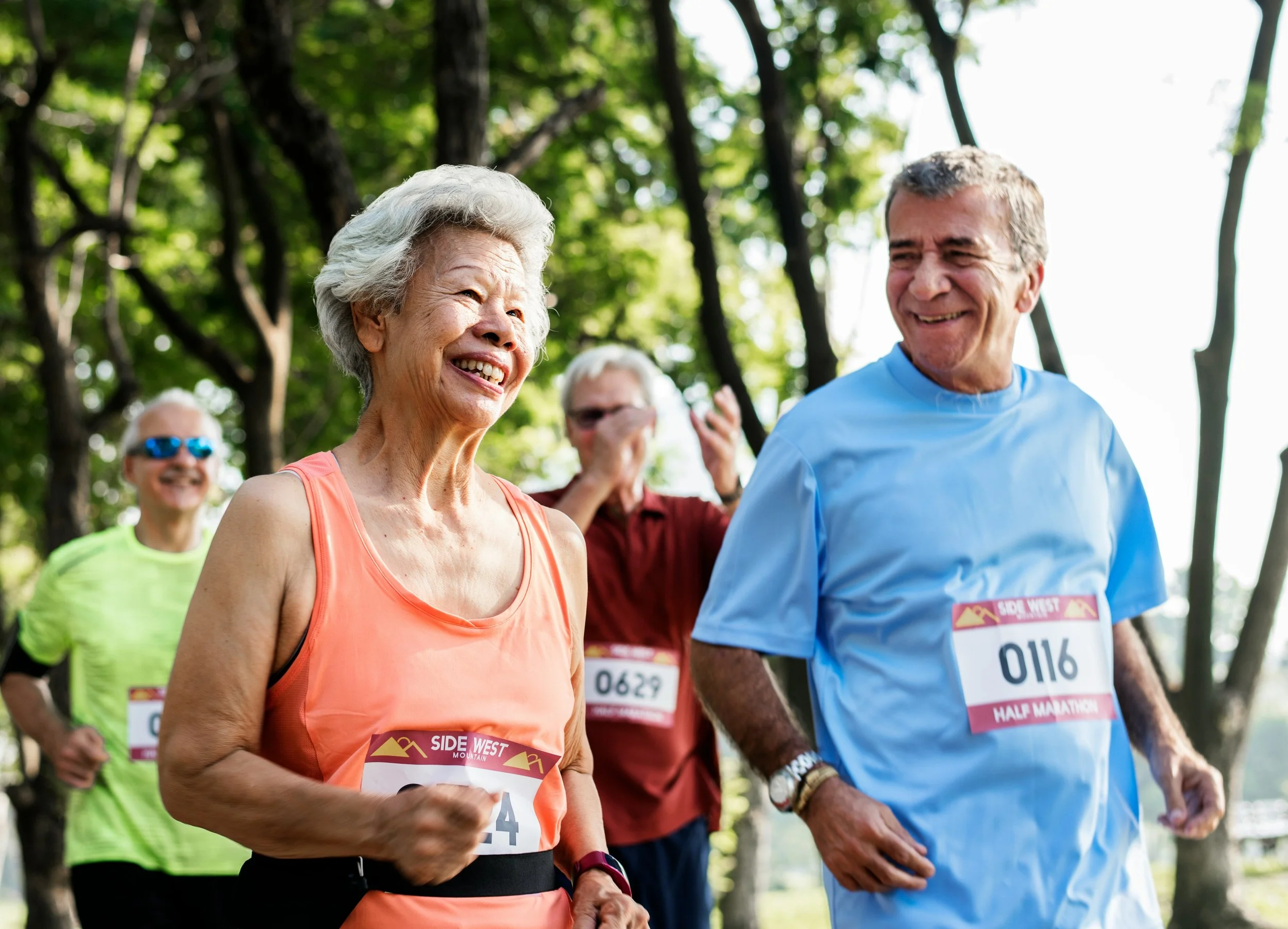 A group of elderly people participating in a half marathon, smiling and running outdoors among trees, wearing running clothes and numbered bibs.
