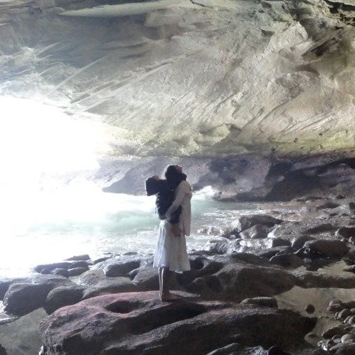A woman with a child on her back standing on rocks inside a cave formation near the ocean, with sunlight streaming in from an opening.