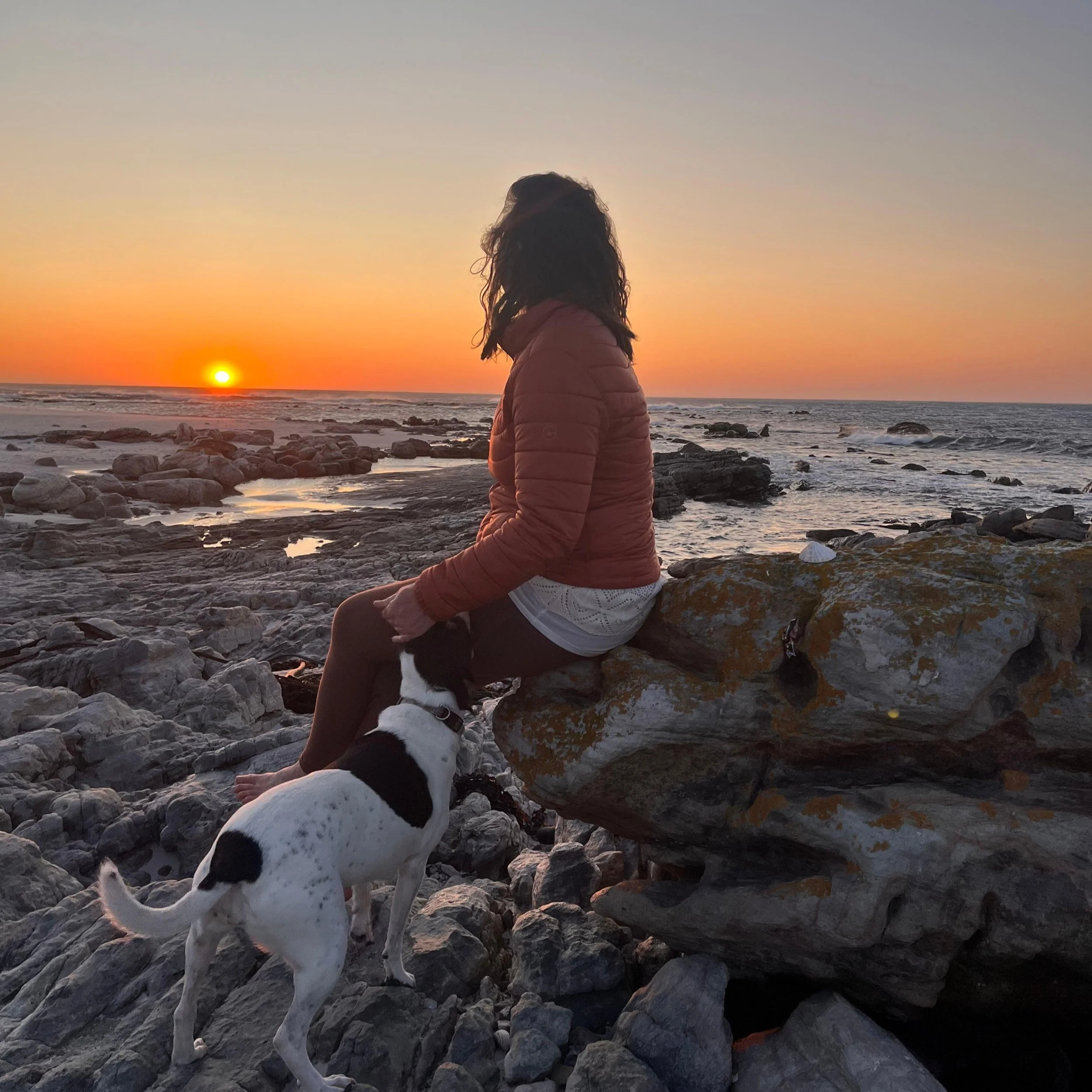 A woman sitting on a large rock by the rocky shoreline at sunset with her dog, watching the sunset over the ocean.