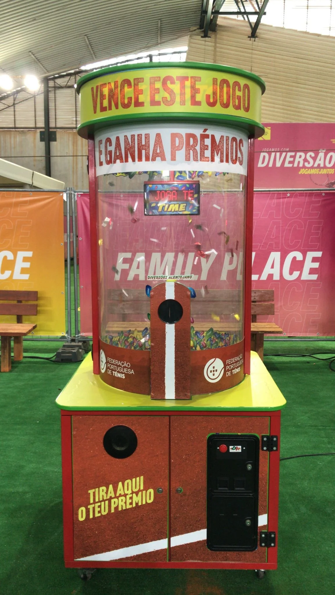 Máquina de caça níquel infantil com letras em português convidando a vencer o jogo, ganhar prêmios e participar, no ambiente de uma feira ou evento de entretenimento, decorada com cores vibrantes e banners de fundo.