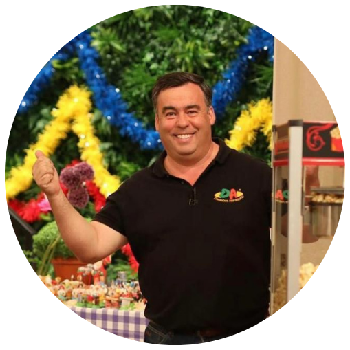 Homem sorridente fazendo sinal de positivo em evento decorado com árvores verdes e enfeites coloridos ao fundo.