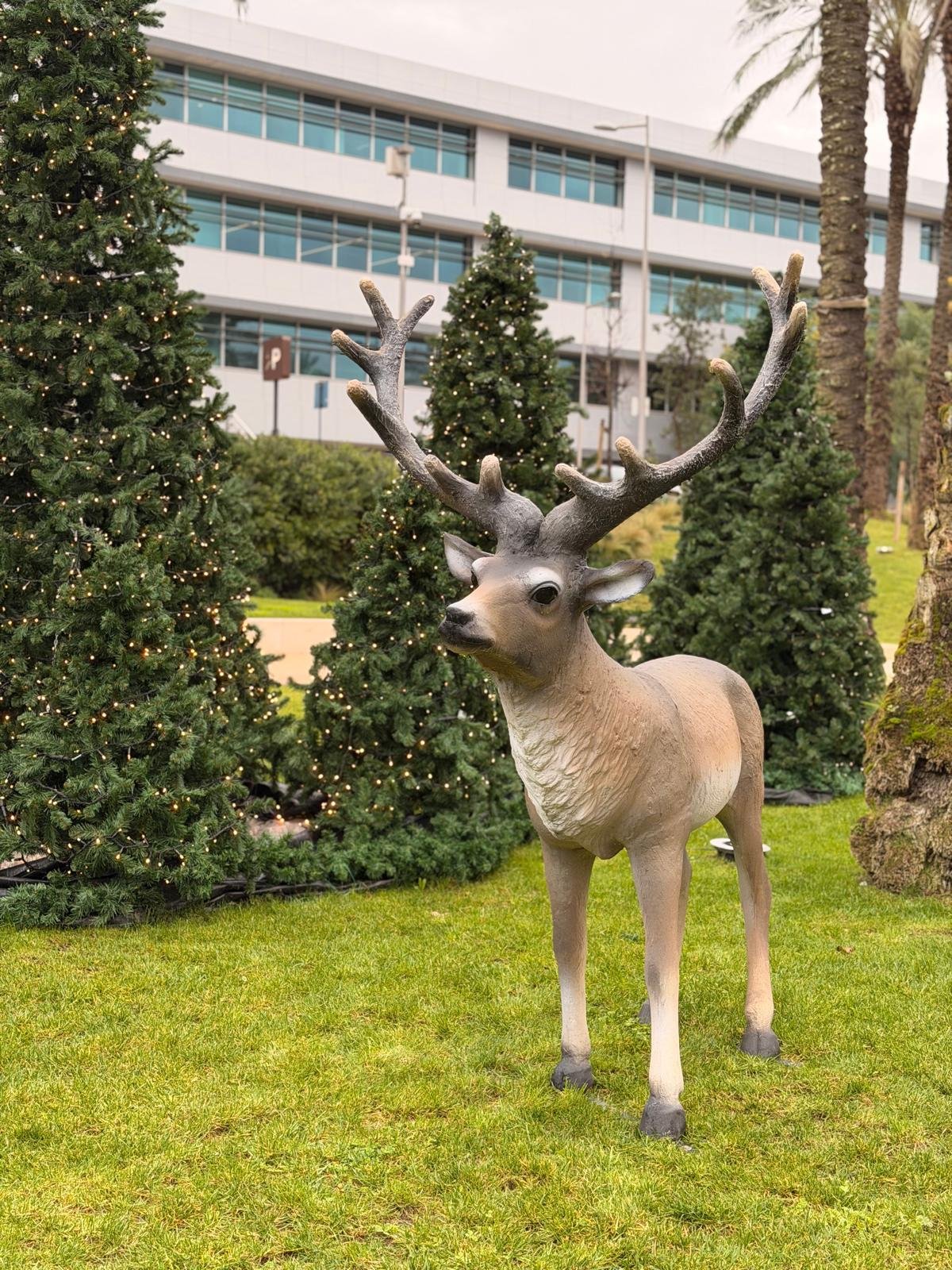 Estátua de um cervo de jardim com ramos de chifres detalhados, cercado por árvores de Natal decoradas com luzes.