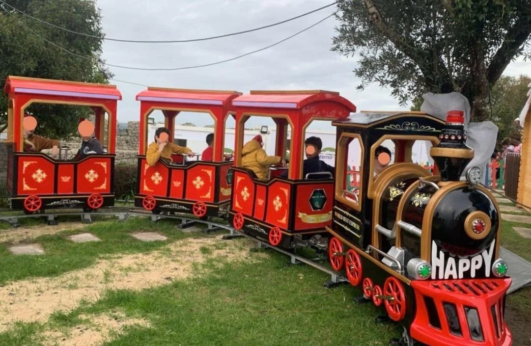 Mini trem de parque com três cabines vermelhas e uma preta, crianças sorrindo e brincando ao ar livre