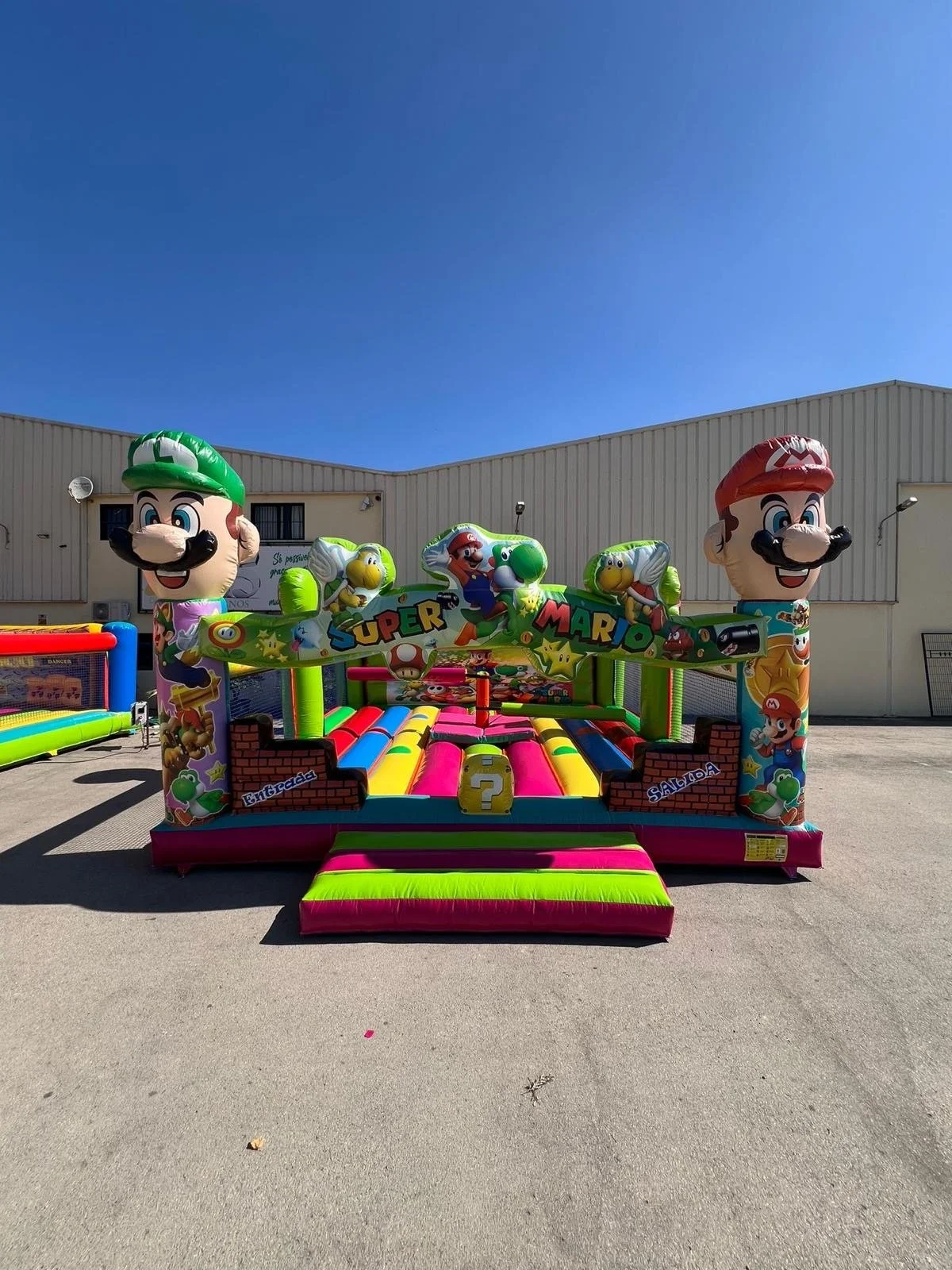 Brinquedo inflável de Mario Bros com cores vibrantes, figuras de Mario, Luigi e Yoshi, em um espaço ao ar livre sob céu azul.