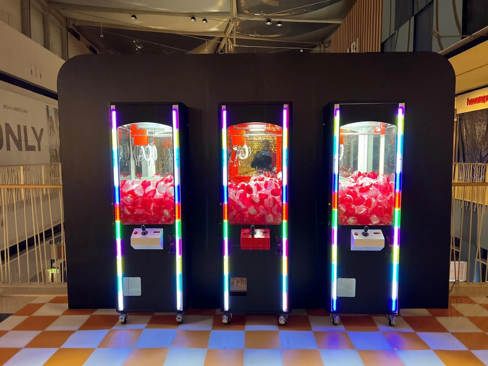 Três máquinas de caça níquel, com luzes coloridas, preenchidas com cubos de plástico em uma área de shopping ou feira.