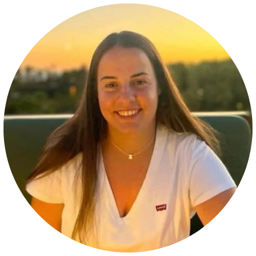 Mulher jovem sorrindo ao pôr do sol, usando camiseta branca e colar, com fundo de céu alaranjado e cenário urbano ao longe.