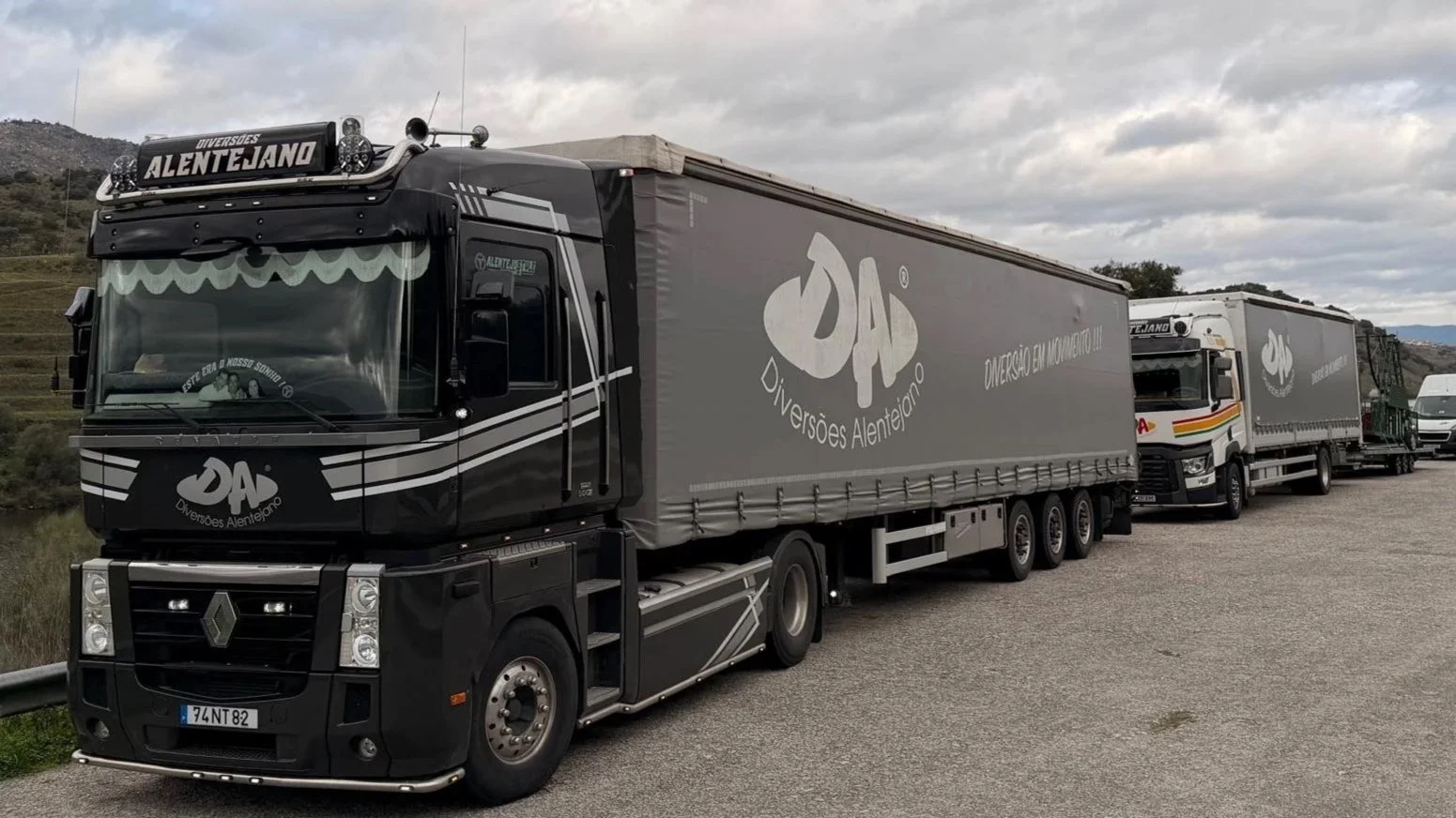Caminhão preto com inscrições e logotipo de Divisões Alentejano, seguido por outros caminhões na estrada