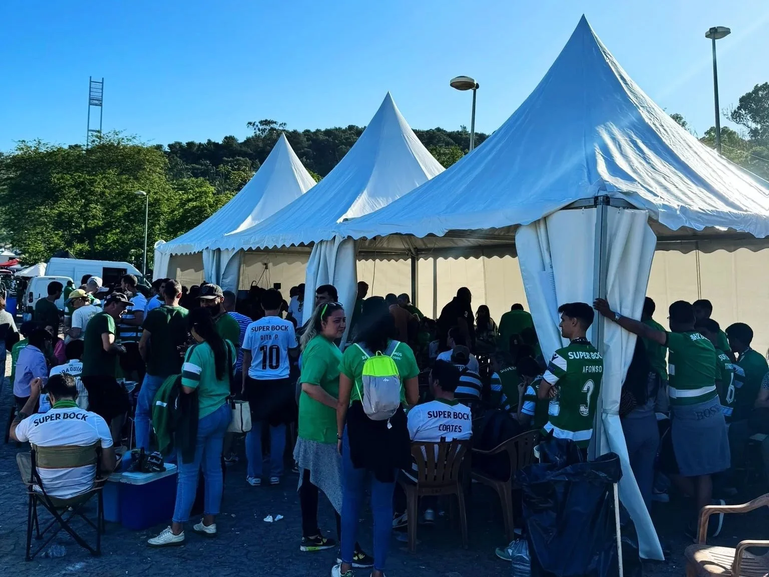 Reunião de pessoas sob uma tendas brancas ao ar livre, com várias pessoas usando camisetas verdes, brancas e pretas, algumas com nomes e números, em um ambiente de evento ou feira.