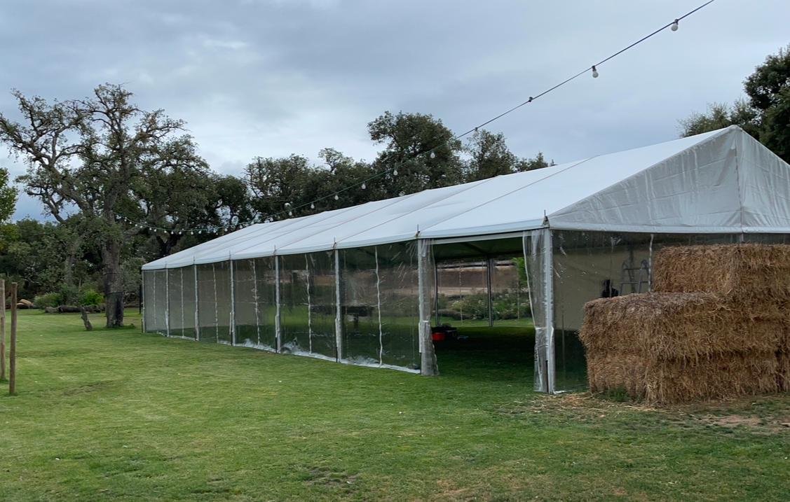 tenda grande com laterais de plástico transparente em um campo gramado com árvores ao fundo e fardos de feno ao lado