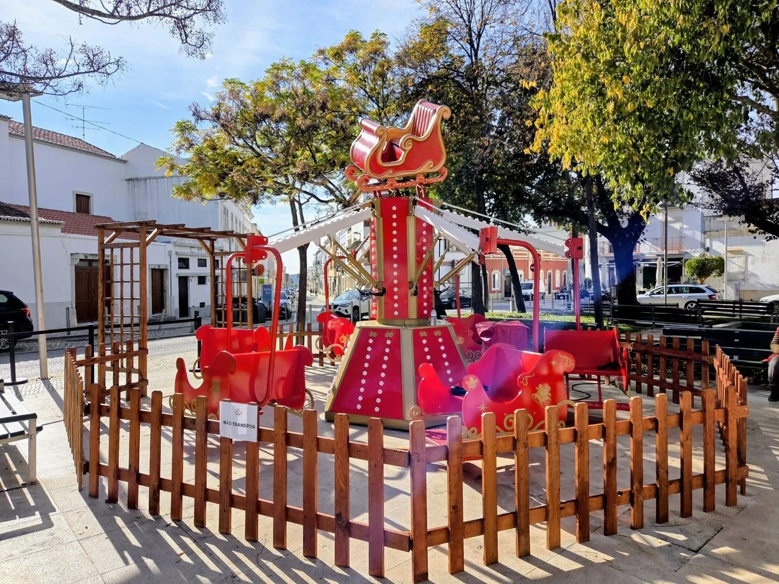 Parque infantil com carruagens de cavalos de brinquedo, cercado por cerca de madeira, em um espaço ao ar livre com árvores e edifícios ao fundo.