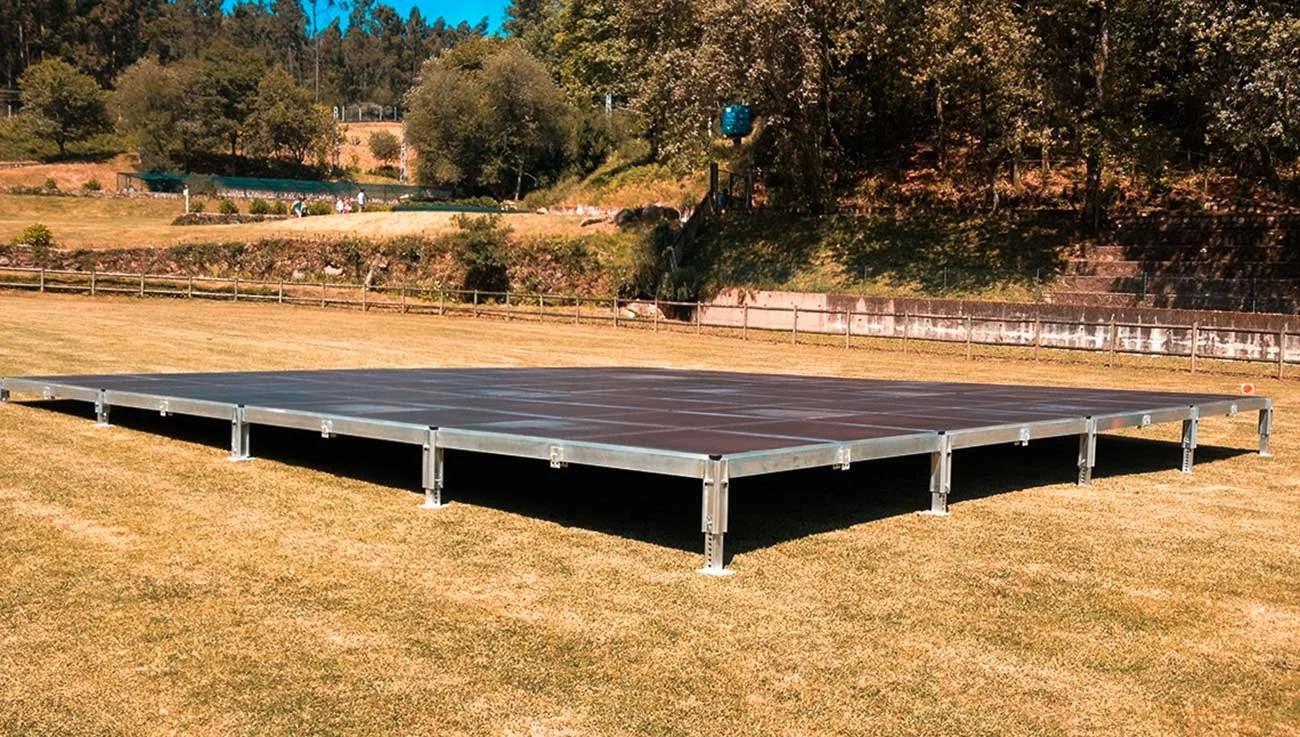 Uma quadra de tênis ao ar livre, elevada do solo com estrutura de metal, cercada por grama e árvores ao fundo.