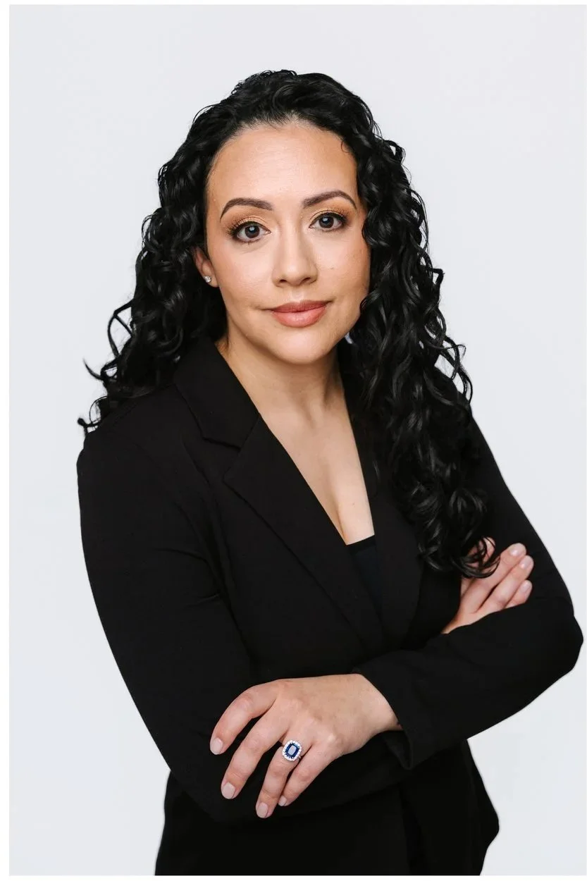 A woman with long, curly black hair dressed in a black blazer, looking confidently at the camera with arms crossed and wearing a ring with a large blue gemstone. Karla Murphy by Storm Pierre Photography in Jacksonville NC Branding Photos